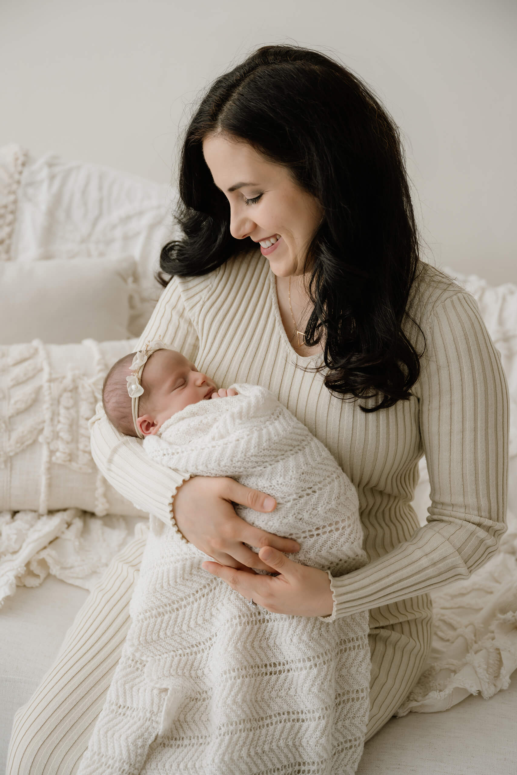 Parent smiles down while holding a newborn wrapped in a white knit blanket on a cream couch.
