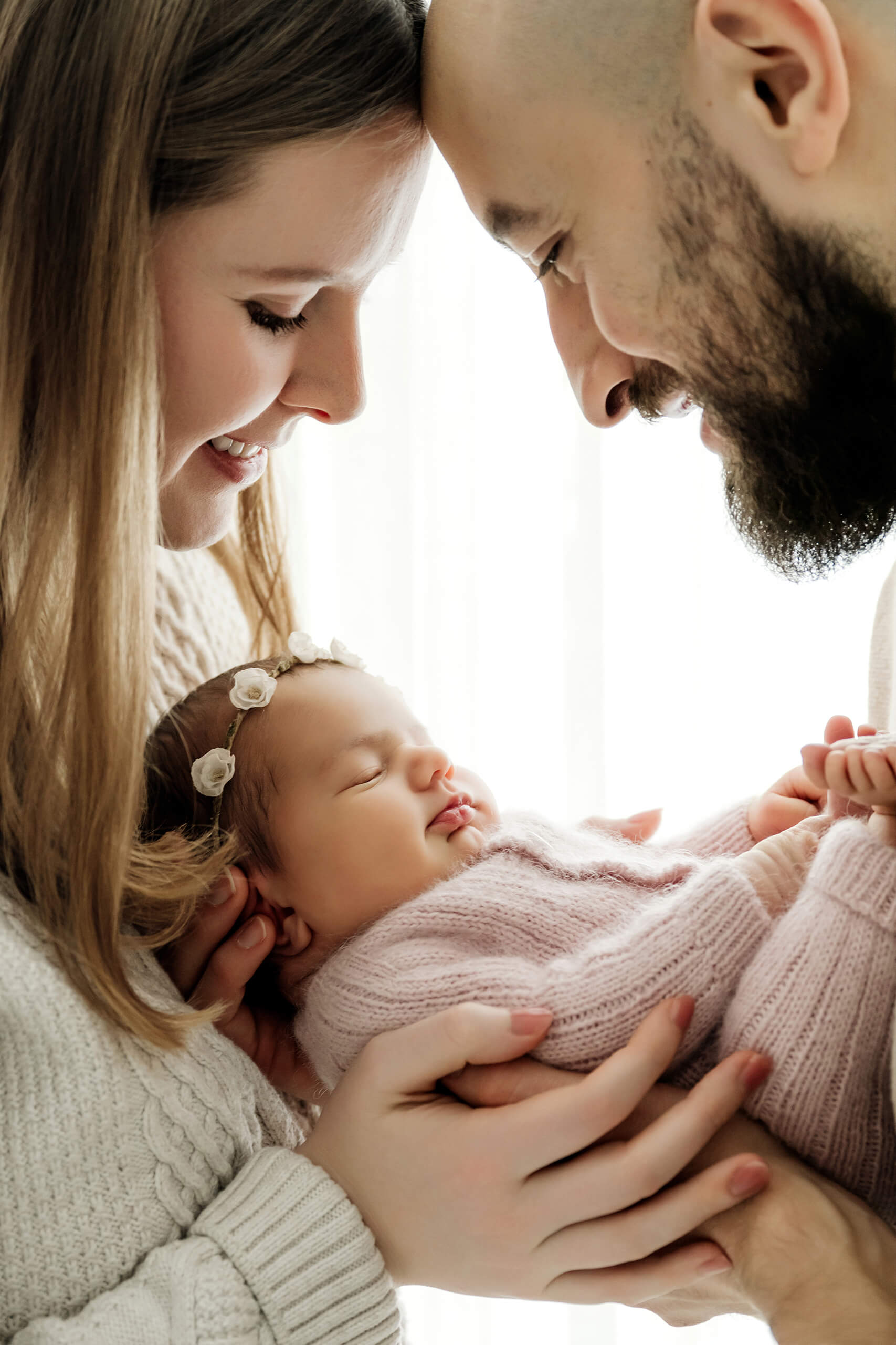 Two parents lean close over their newborn held between them in a pale pink knit outfit.
