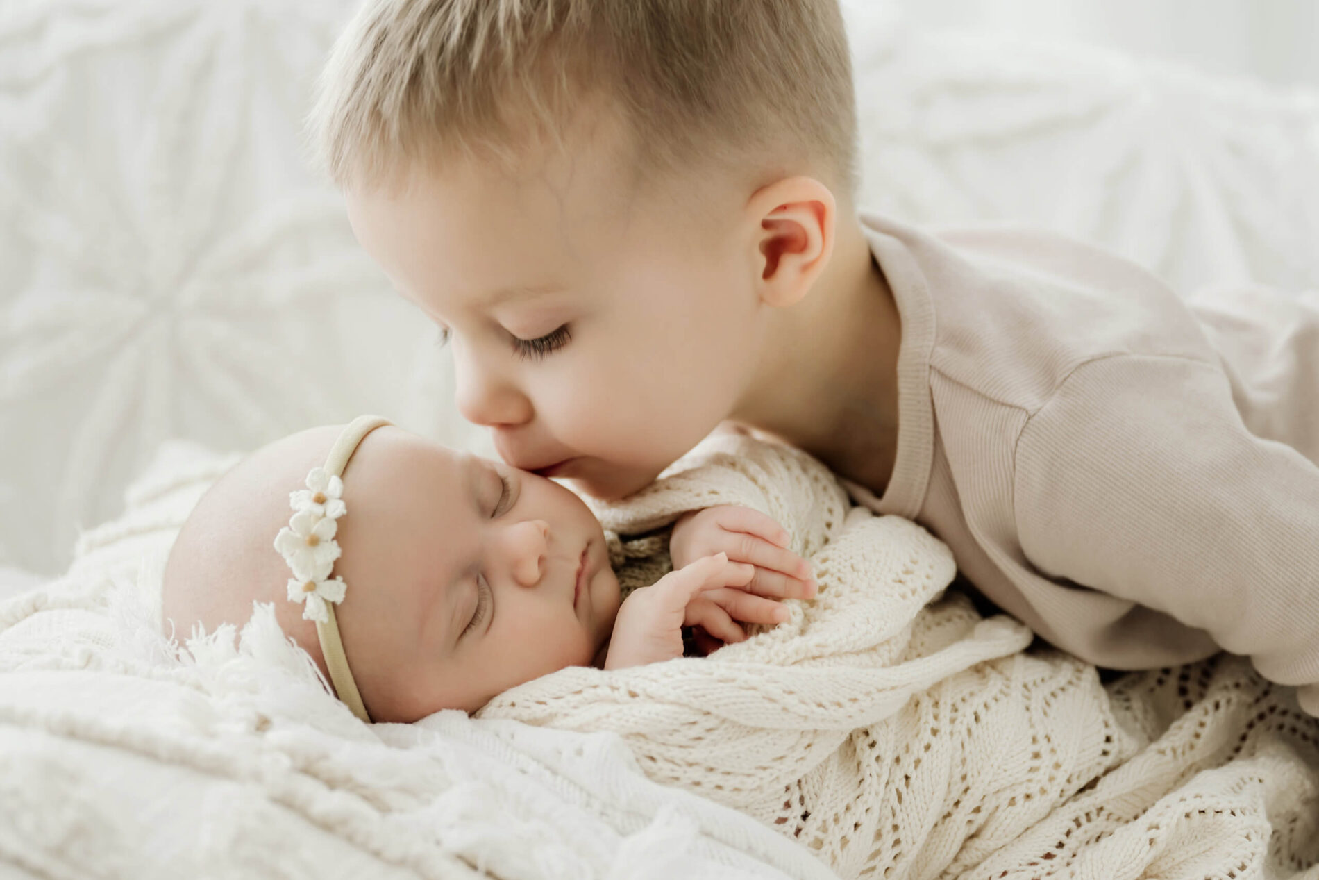 Young sibling kisses a newborn with eyes closed on a cream couch.