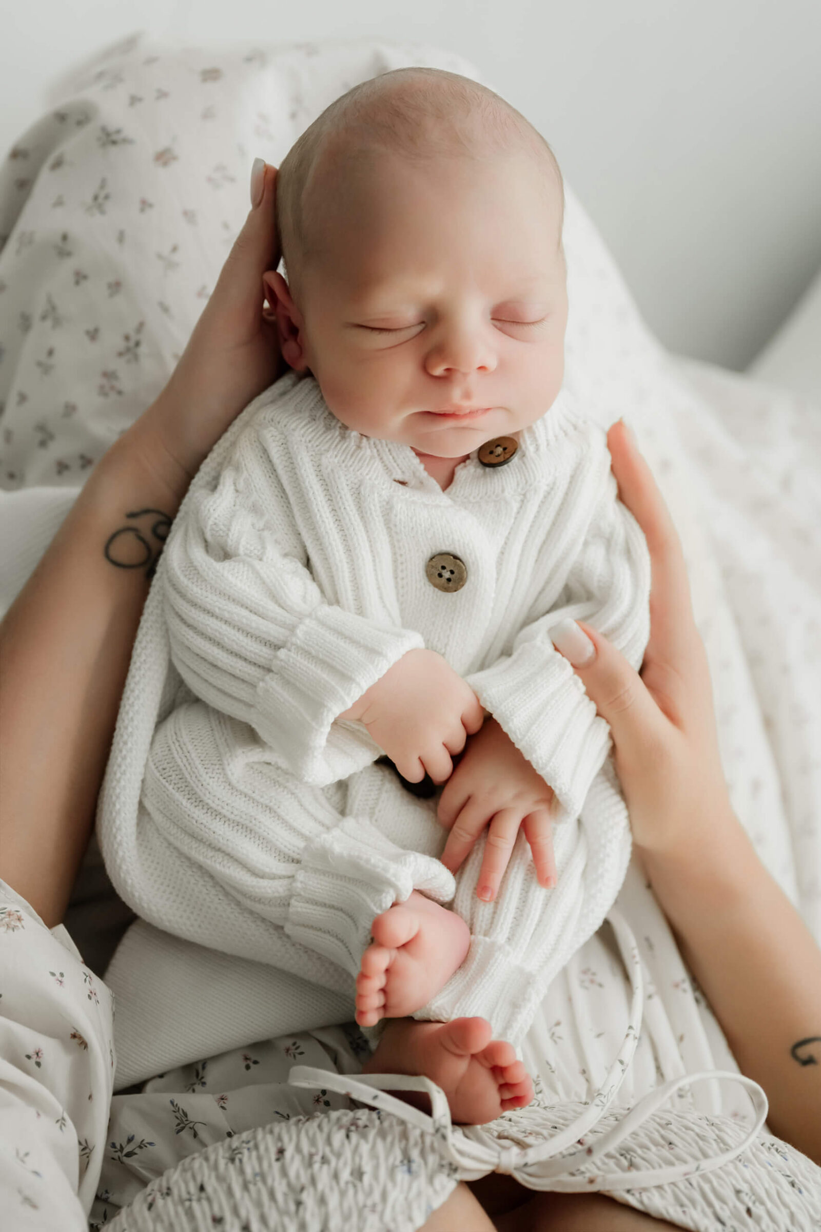 Newborn with eyes closed sits supported in an adult’s lap, wearing a white knit outfit with brown buttons.