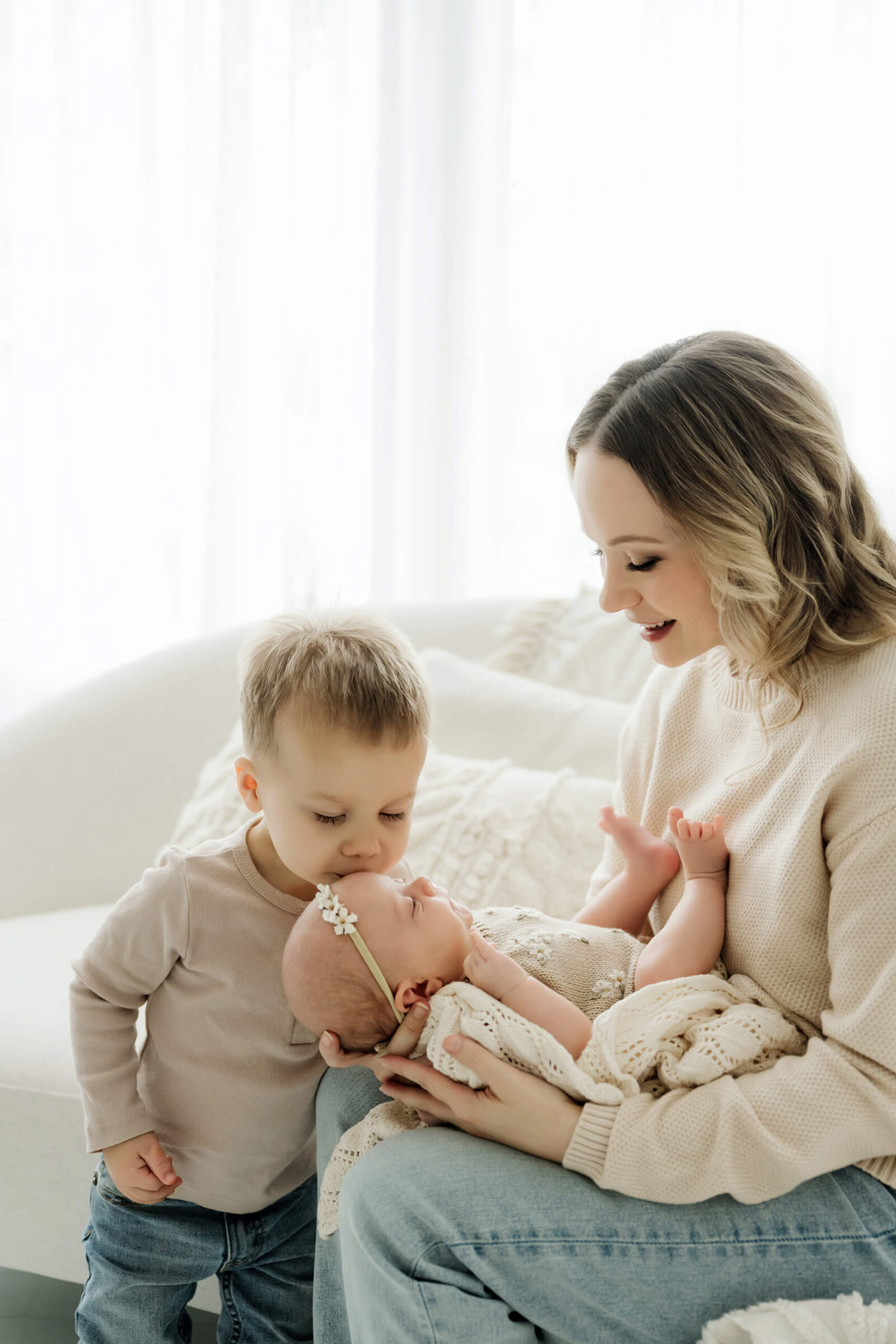 Young sibling kisses a newborn’s cheek while an adult cradles the baby in a cream knit blanket.
