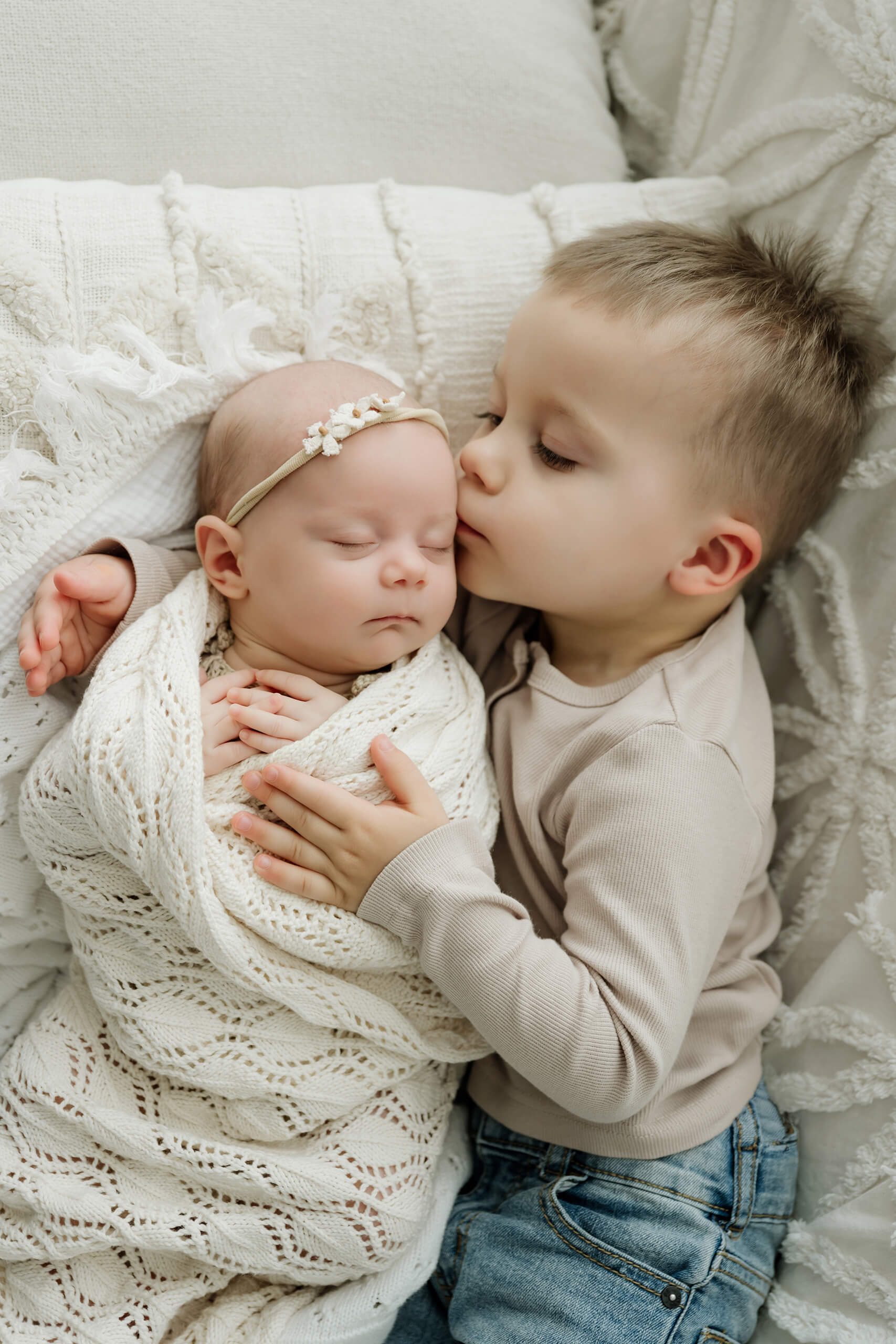 Young sibling kisses a newborn’s forehead while lying on a cream couch