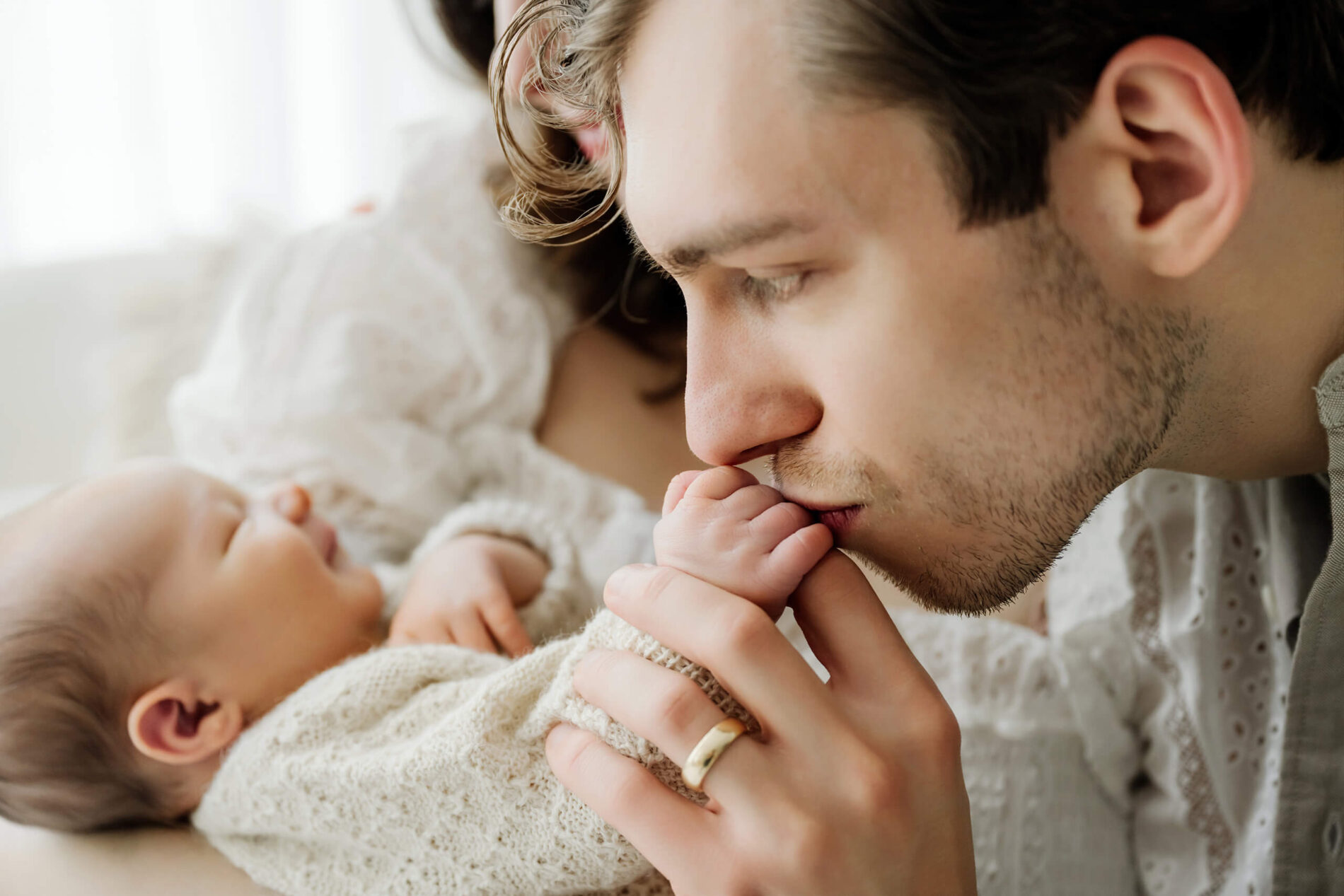 Father kisses his newborn baby’s hand while the baby rests in the mother’s arms.
