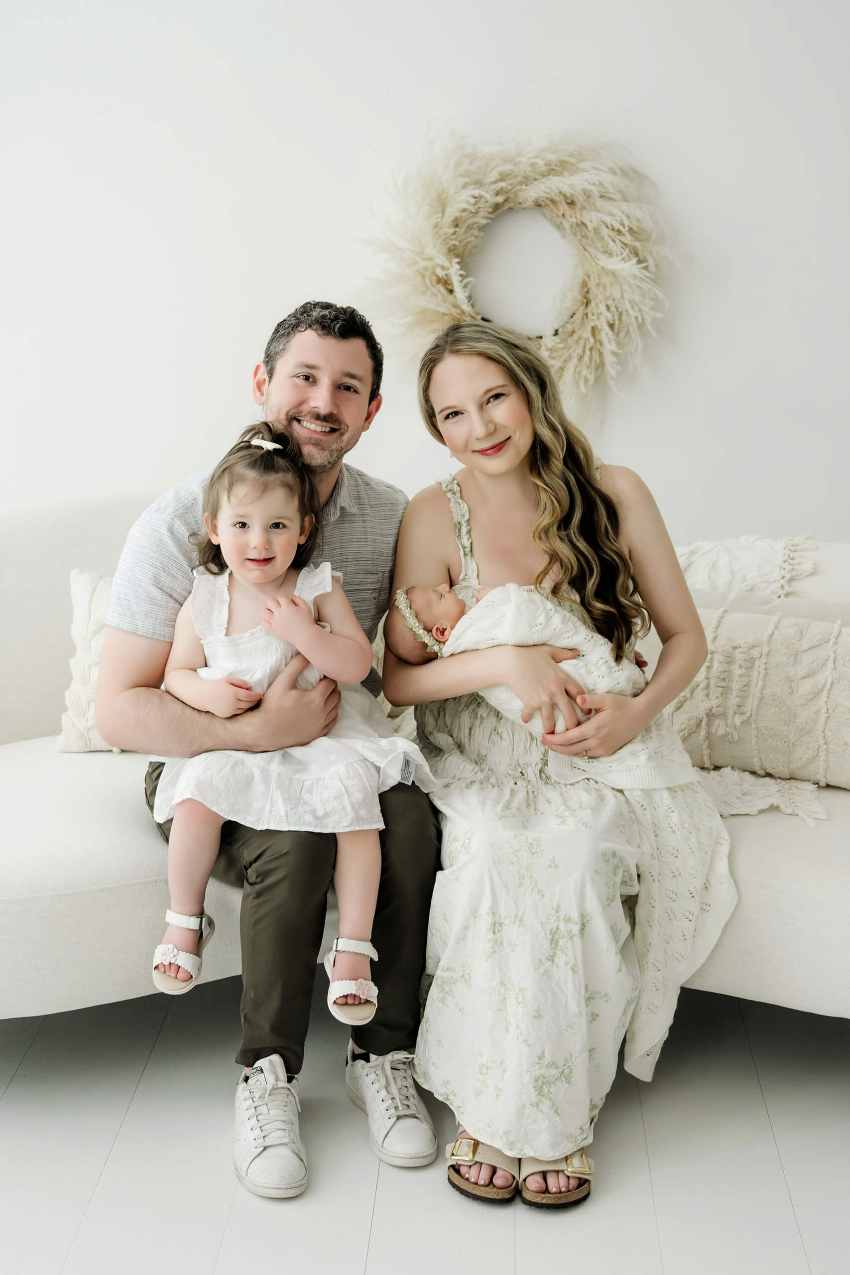 Family of four with parents, toddler daughter, and newborn seated together on a white studio sofa.