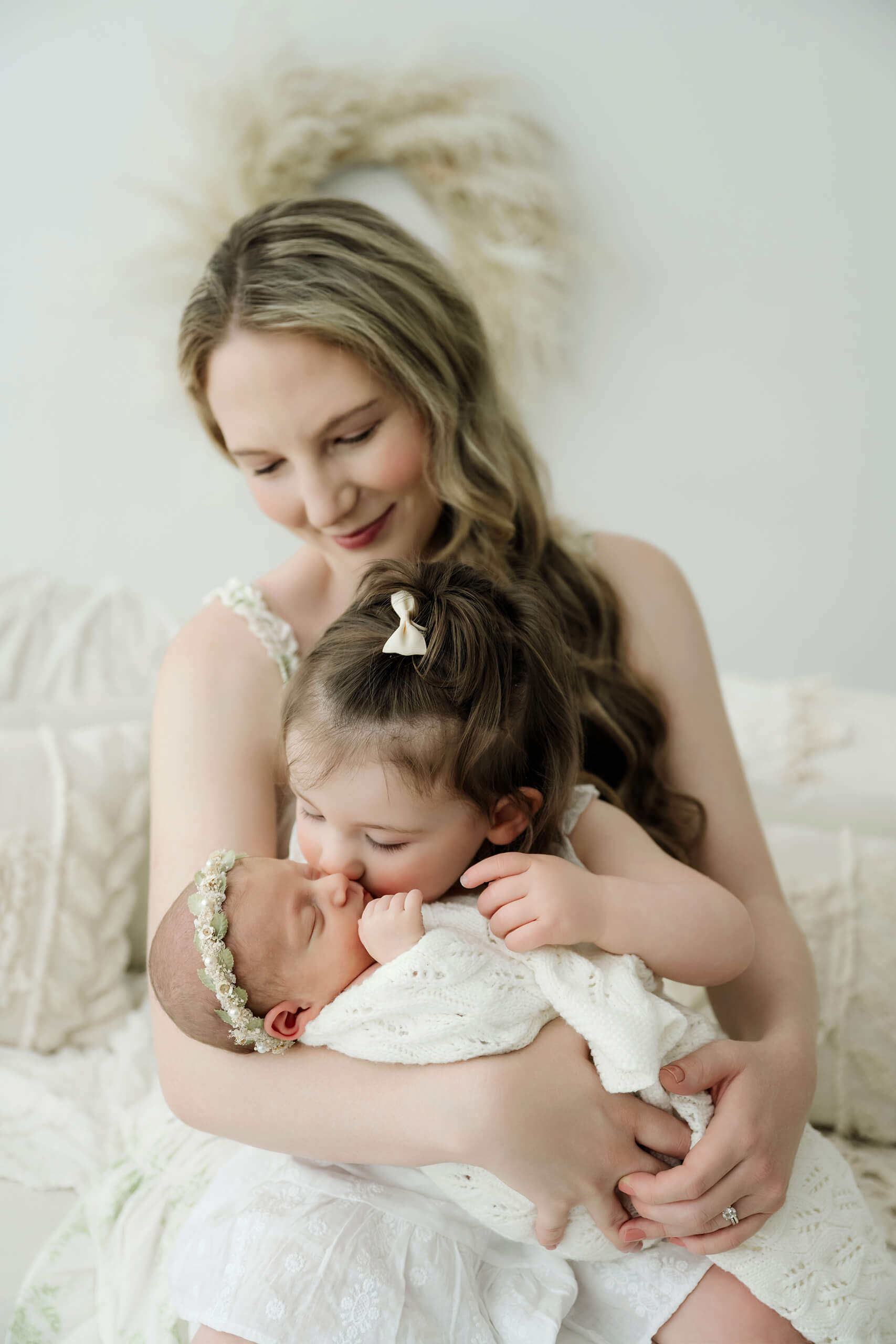 Toddler sister kisses the newborn while their mother holds both children in her arms.