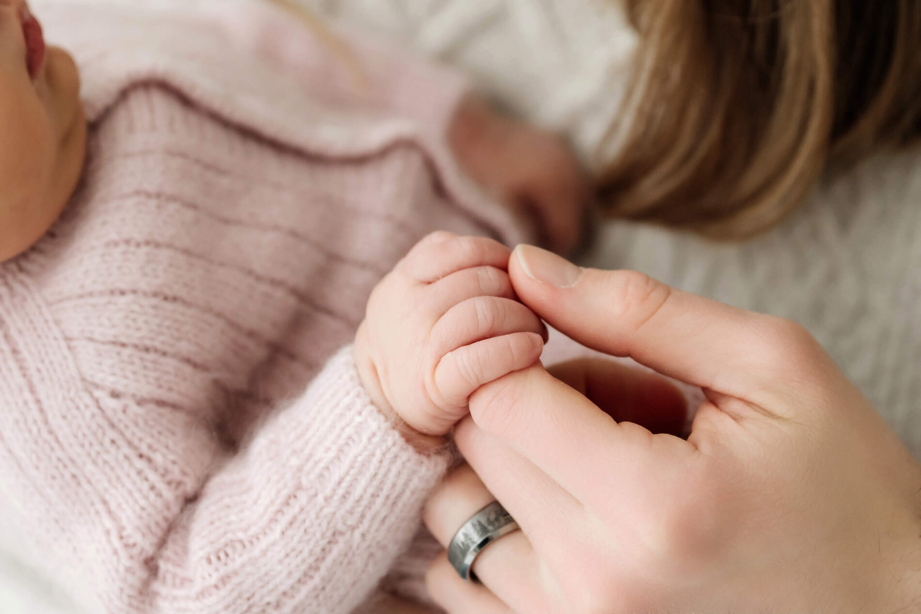 Close-up of a newborn’s hand wrapped around a parent’s finger beside a soft pink knit outfit.