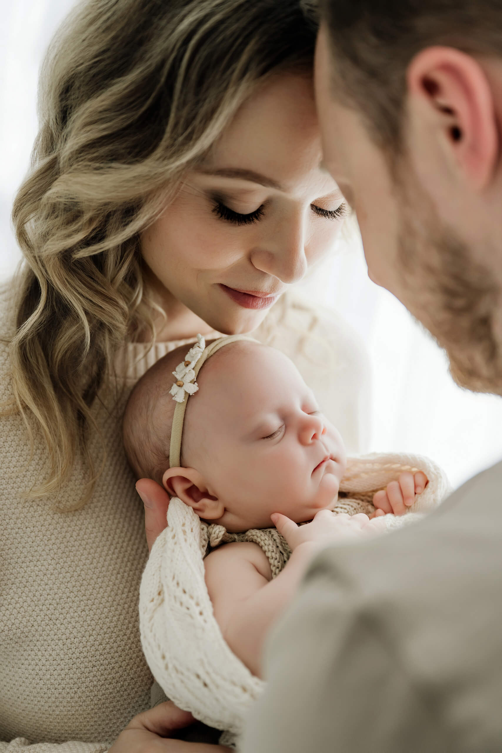 Mother and father lean close around their sleeping newborn in a cream knit outfit and floral headband.