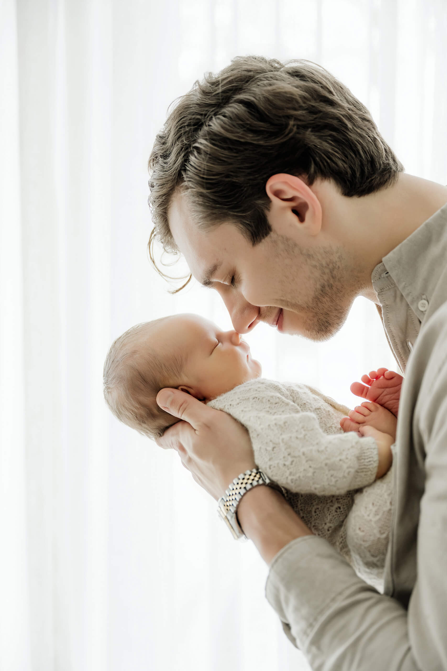 Father gently touches noses with his sleeping newborn while holding the baby near a bright studio window.