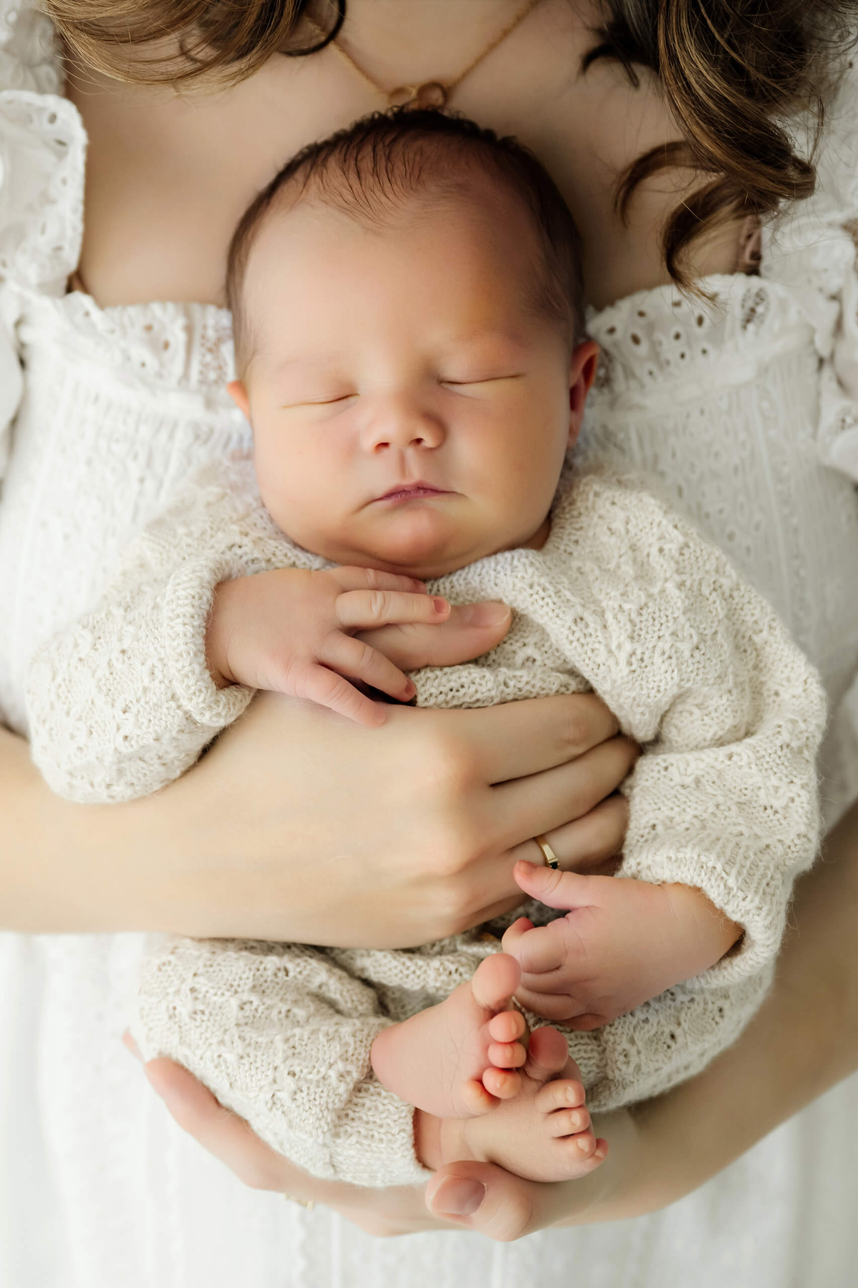 Sleeping newborn in a cream knit outfit rests in the mother’s arms against her white eyelet dress.