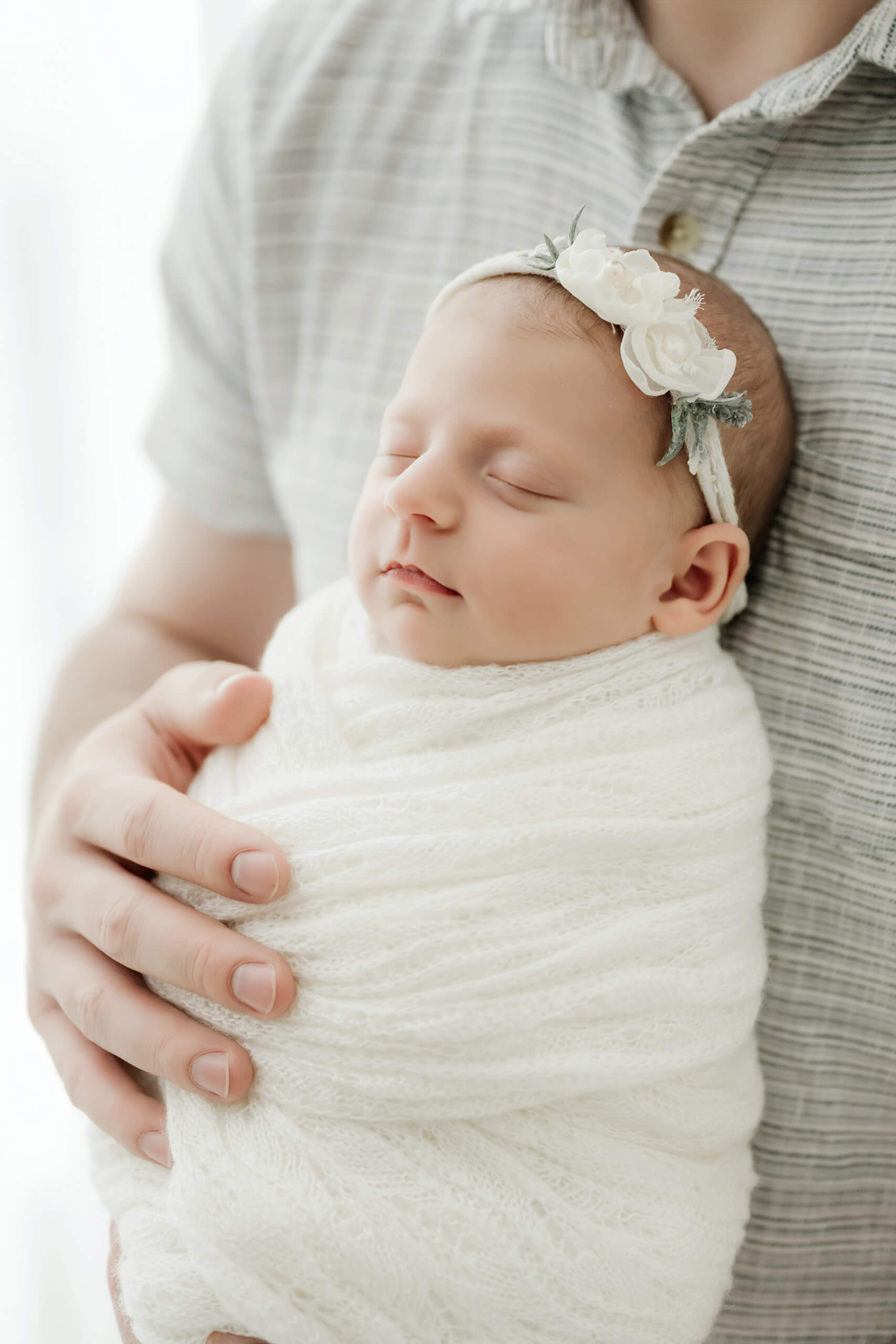 Sleeping newborn wrapped in white and wearing a floral headband rests against the father’s chest.