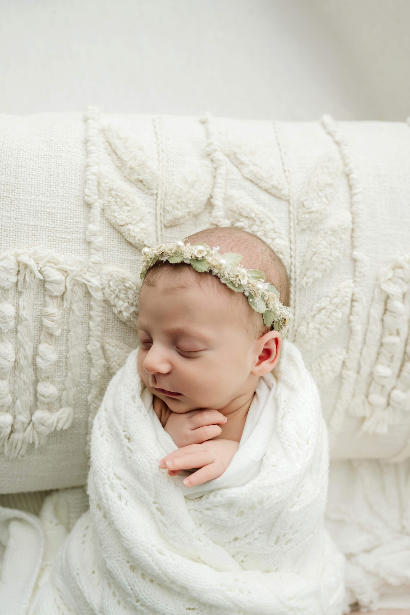 Sleeping newborn wrapped in a white knit blanket and wearing a green floral crown on a textured studio sofa.