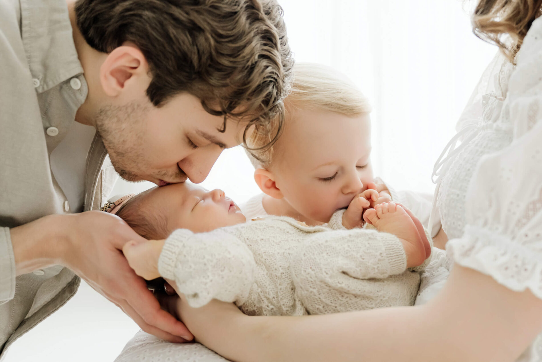 Father kisses his sleeping newborn’s forehead while an older sibling kisses the baby’s foot in the mother’s arms.