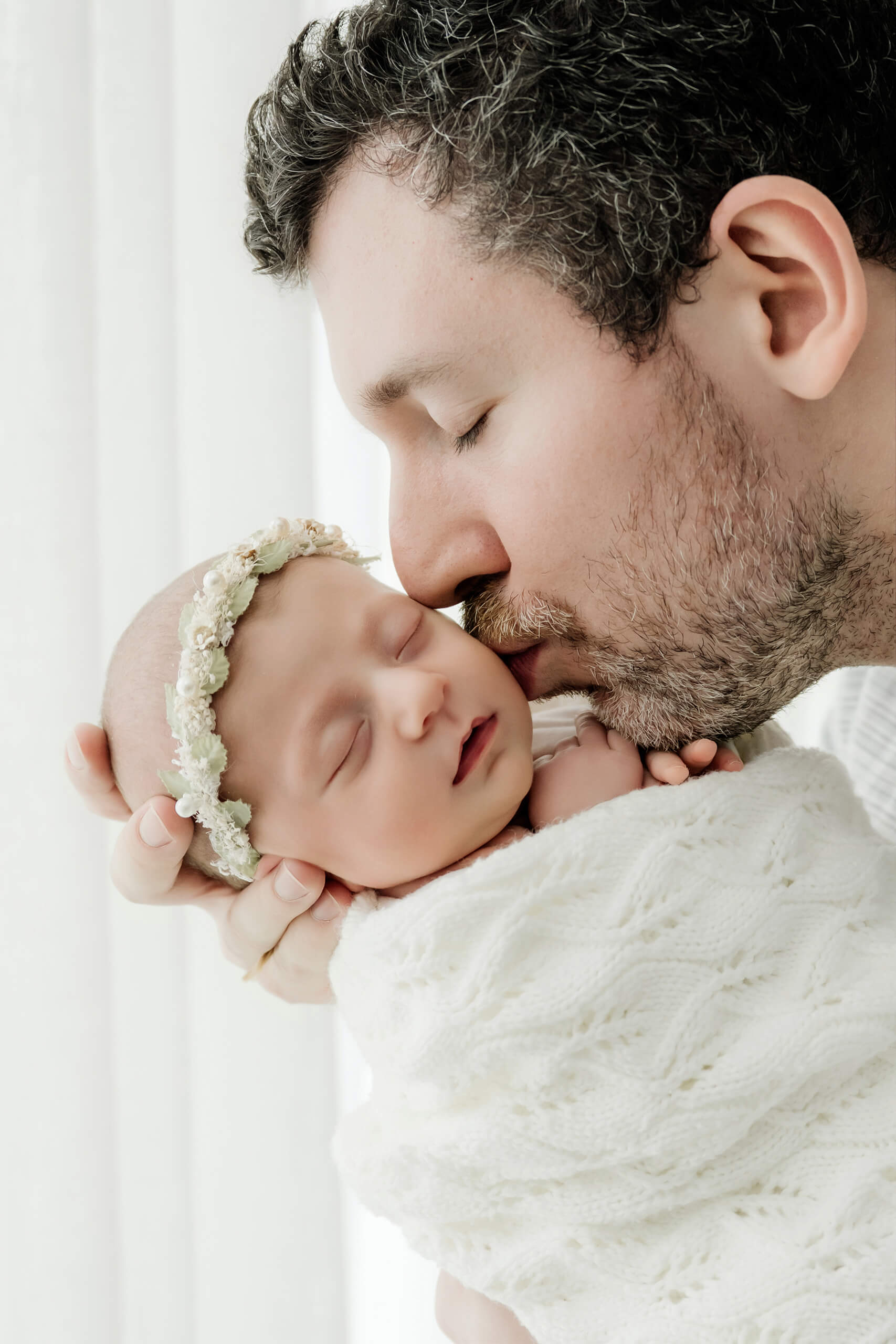 Father kisses his sleeping newborn’s forehead while supporting the baby’s head in his hands.