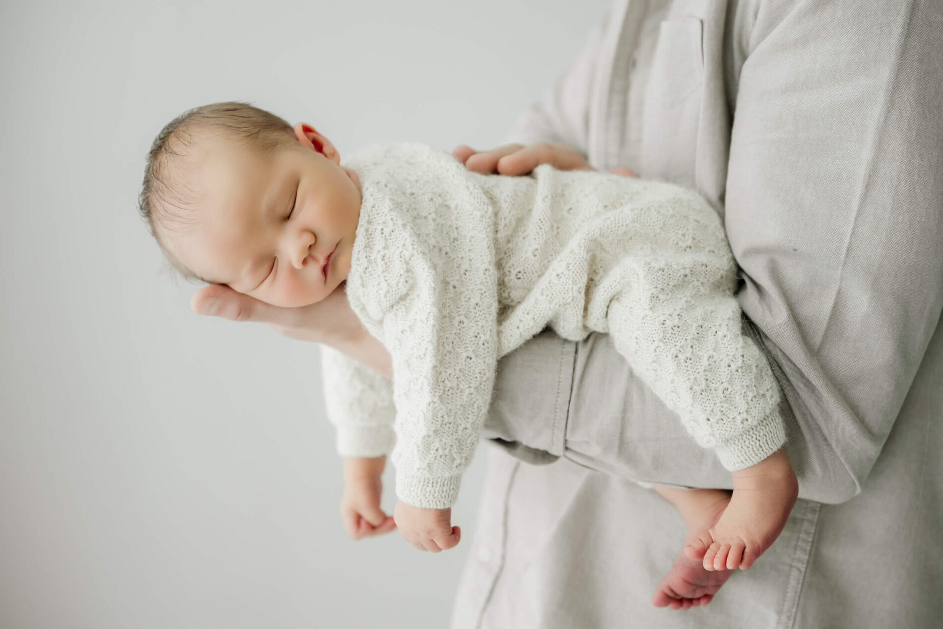 Sleeping newborn in a cream knit outfit rests across a parent’s forearm.
