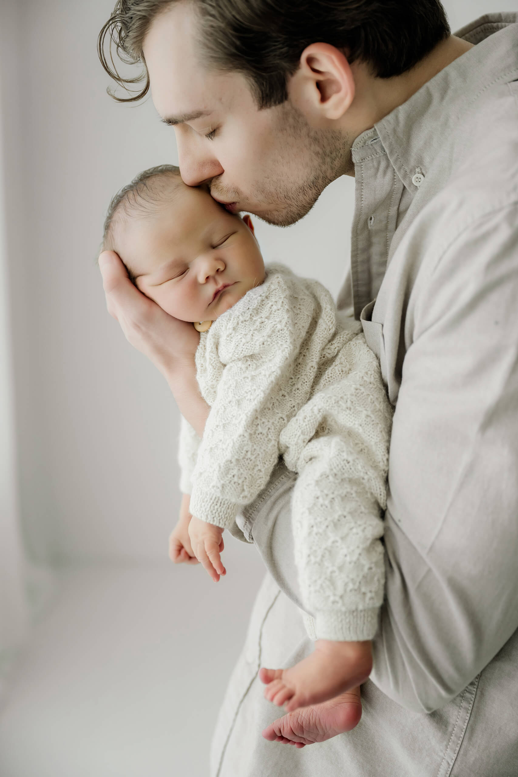 Father kisses his sleeping newborn’s head while holding the baby against his chest.