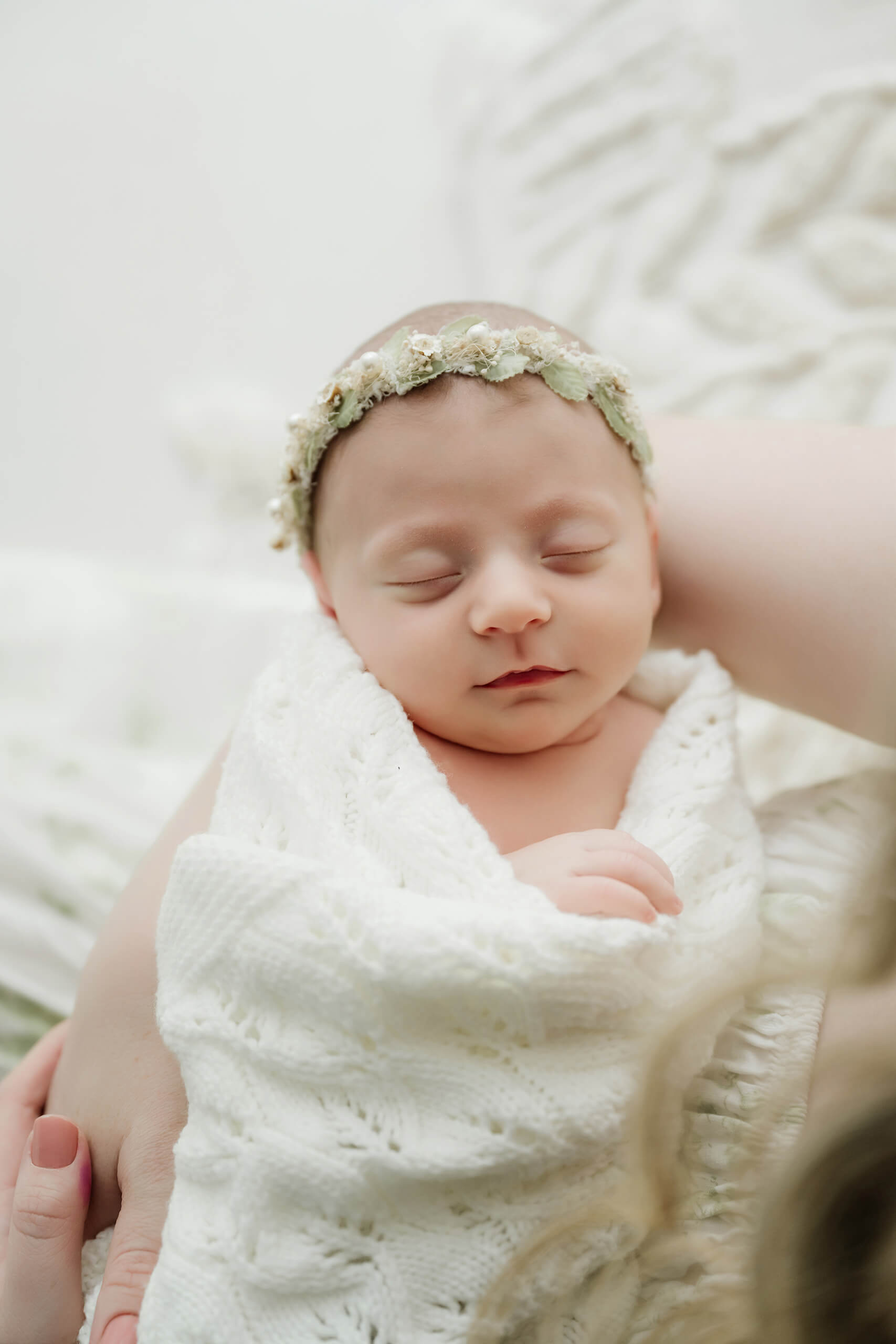 Sleeping newborn wearing a delicate floral crown rests wrapped in a white knit blanket in the mother’s arms.
