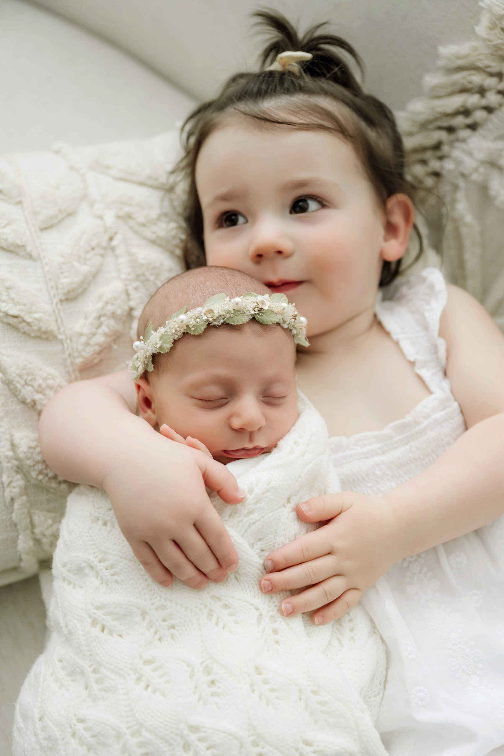 Toddler sister gently holds her sleeping newborn sibling wrapped in white and wearing a floral crown.