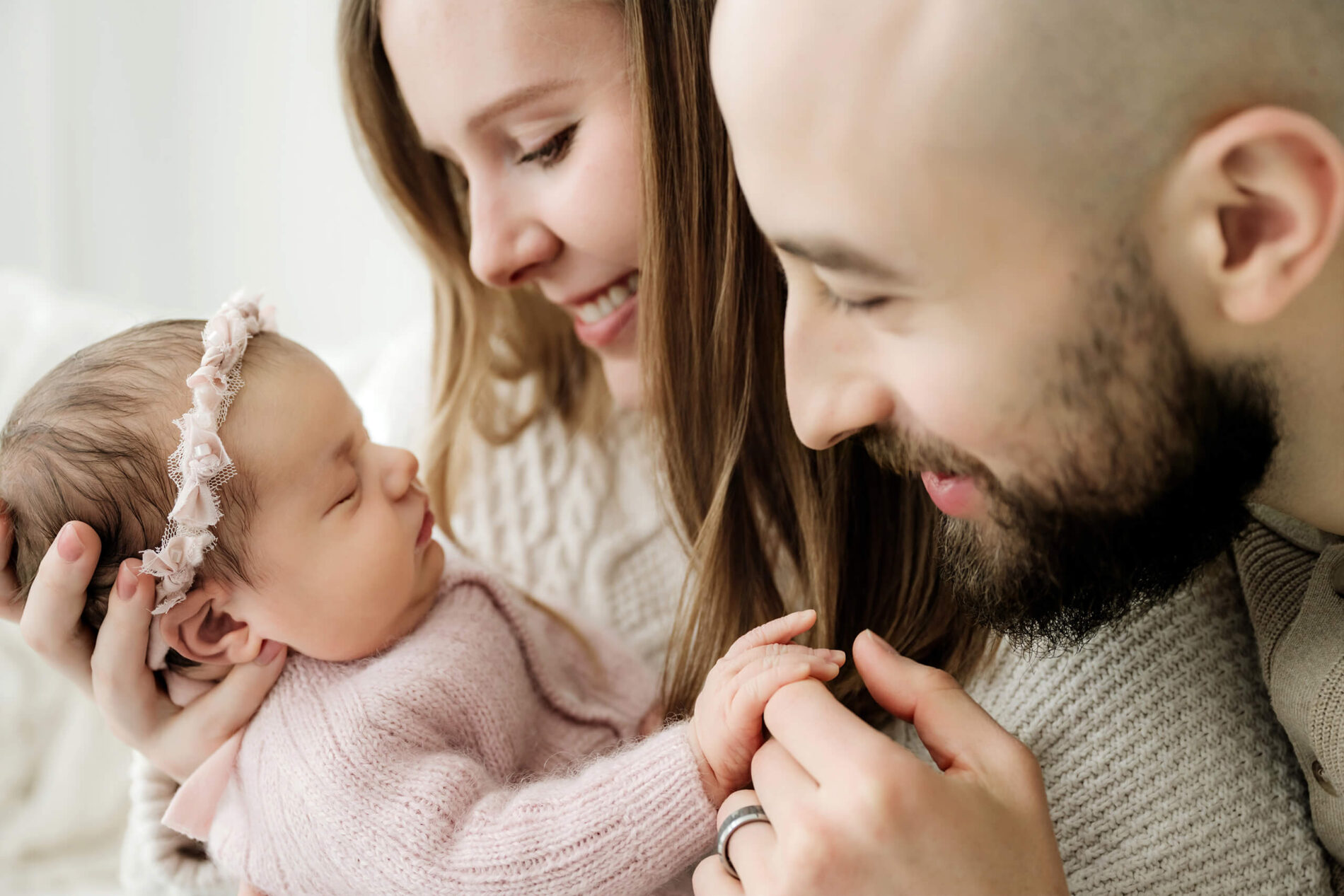 Mother and father smile at their sleeping newborn in a pink knit outfit while holding the baby’s hand.