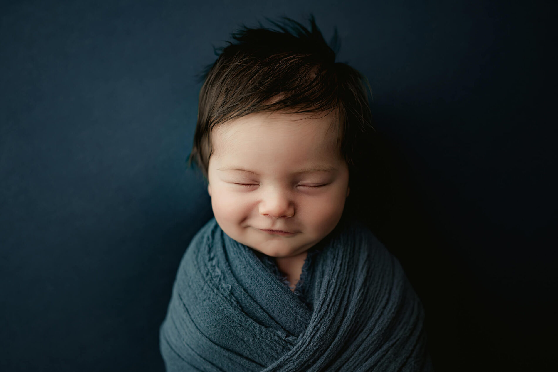 Sleeping newborn with dark hair and a slight smile, wrapped in deep blue against a matching blue backdrop.