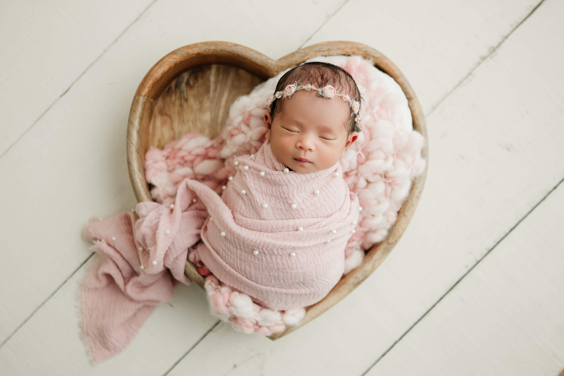 Newborn wrapped in pink fabric with pearl details, resting in a wooden heart-shaped bowl with a floral headband.