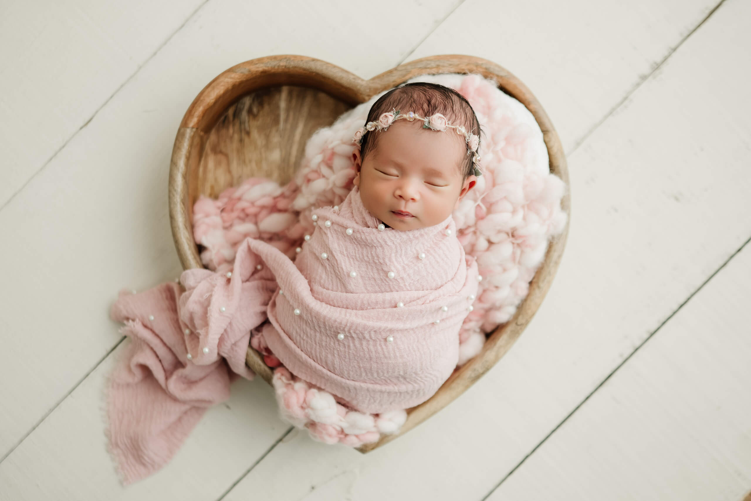 Newborn wrapped in pink fabric with pearl details, resting in a wooden heart-shaped bowl with a floral headband.