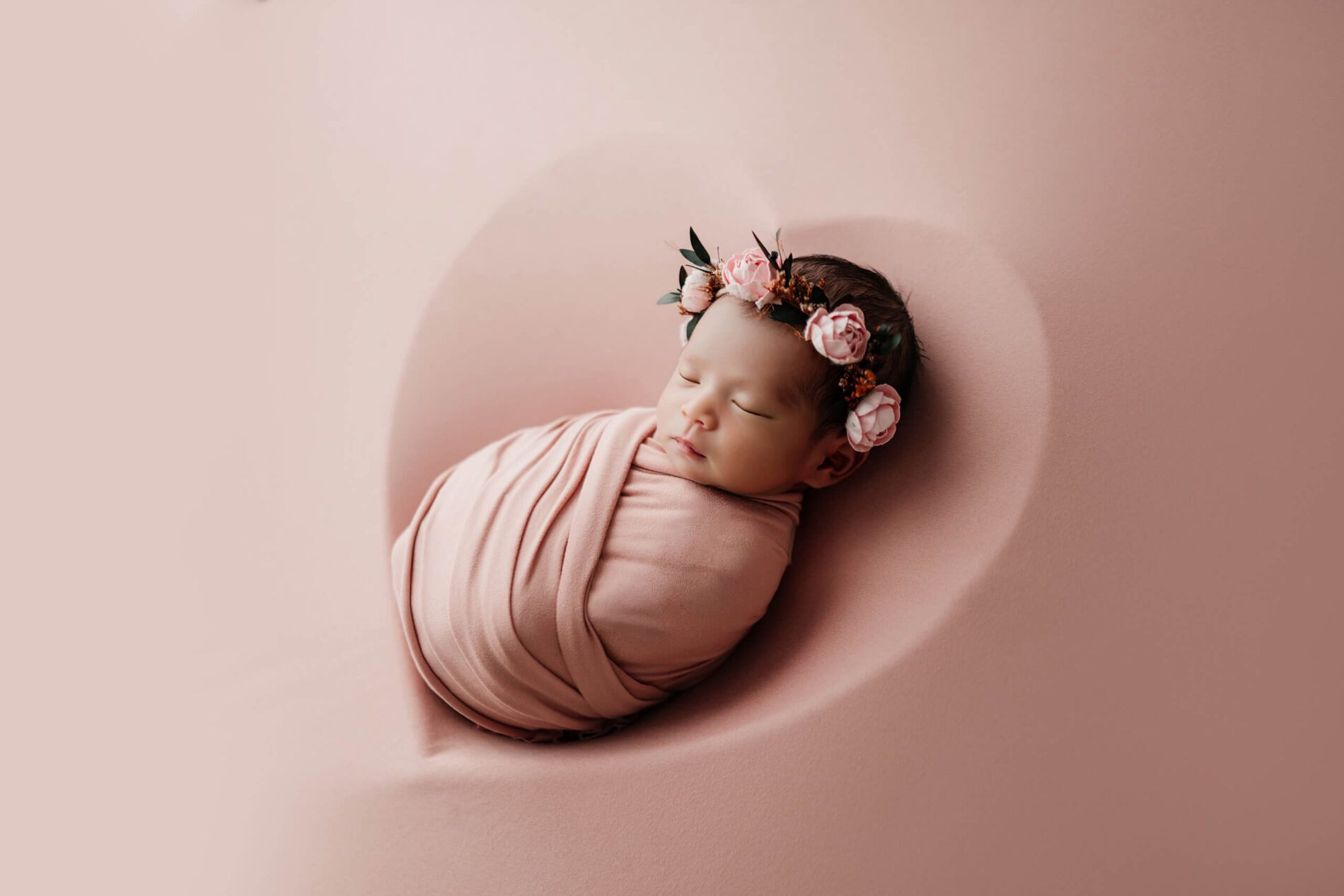 Sleeping newborn wrapped in dusty pink and wearing a floral crown on a matching pink backdrop.