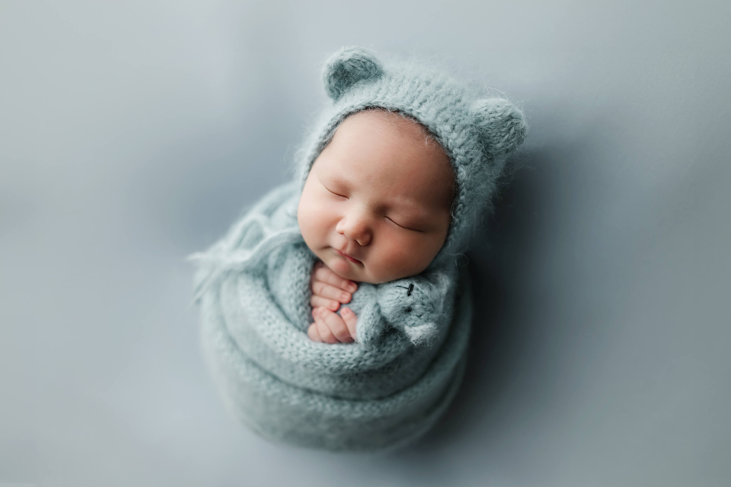 Sleeping newborn wrapped in soft blue knit, wearing a bear-ear bonnet and holding a small matching teddy bear.