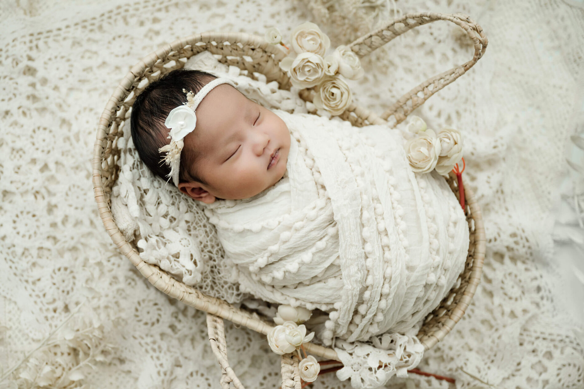 Sleeping newborn wrapped in white and wearing a floral headband, posed in a woven rabbit-shaped basket with cream flowers.