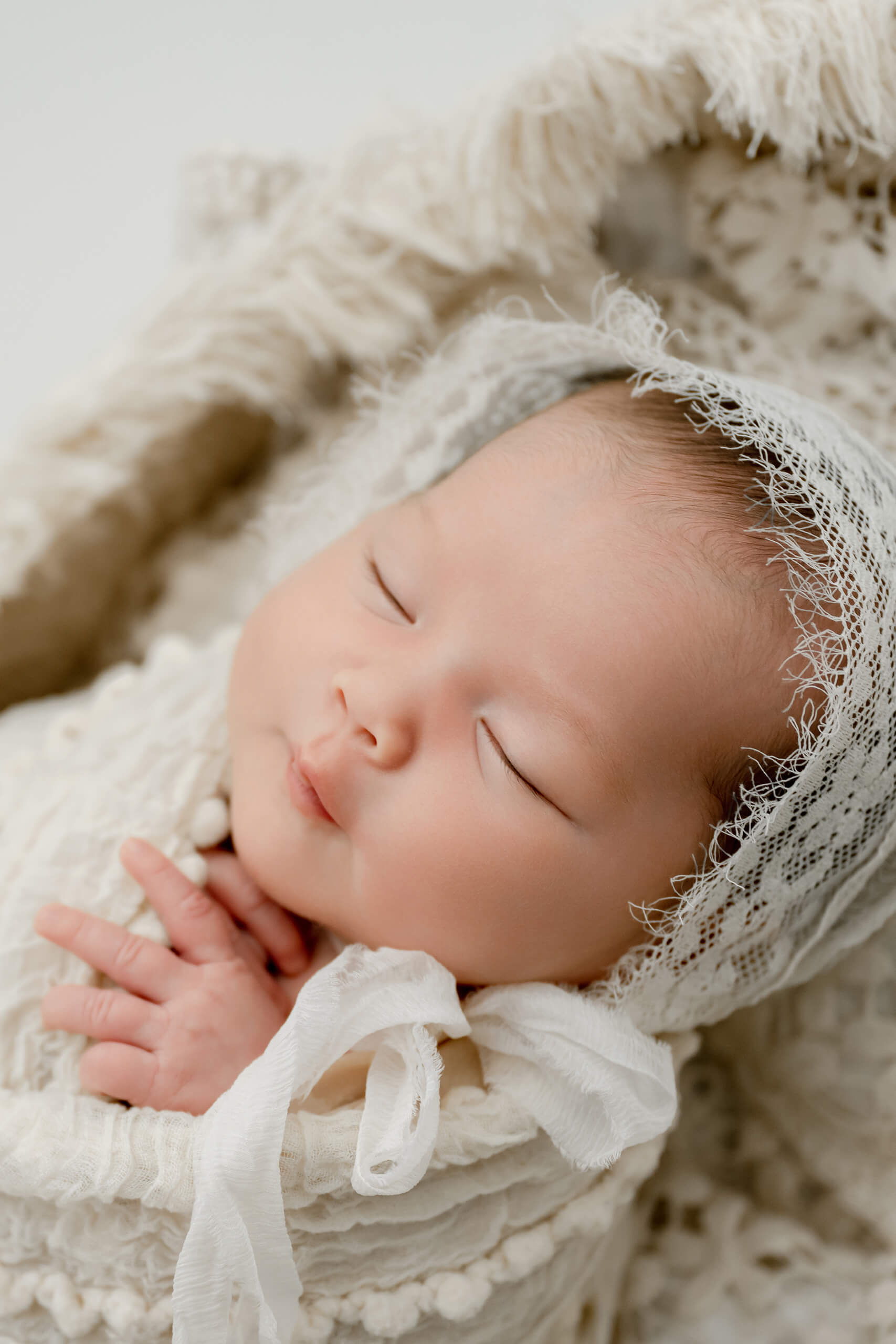 Close-up of a sleeping newborn wrapped in white textured layers and wearing a delicate lace bonnet.