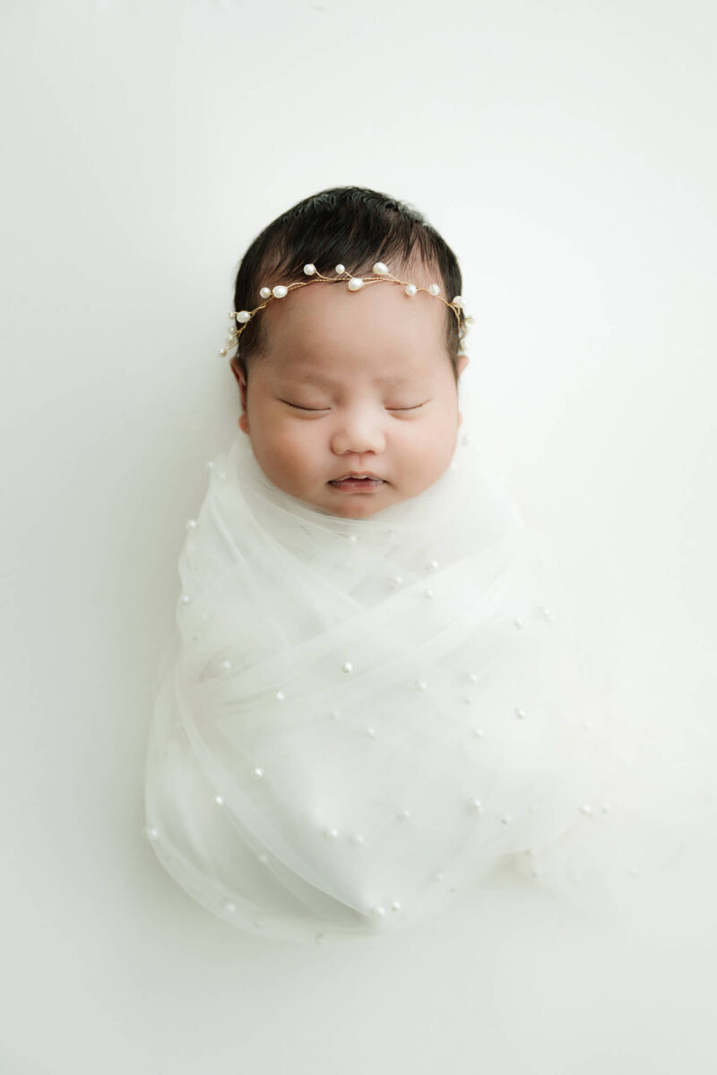 Sleeping newborn wrapped in sheer white fabric with pearl details and wearing a delicate pearl headband.
