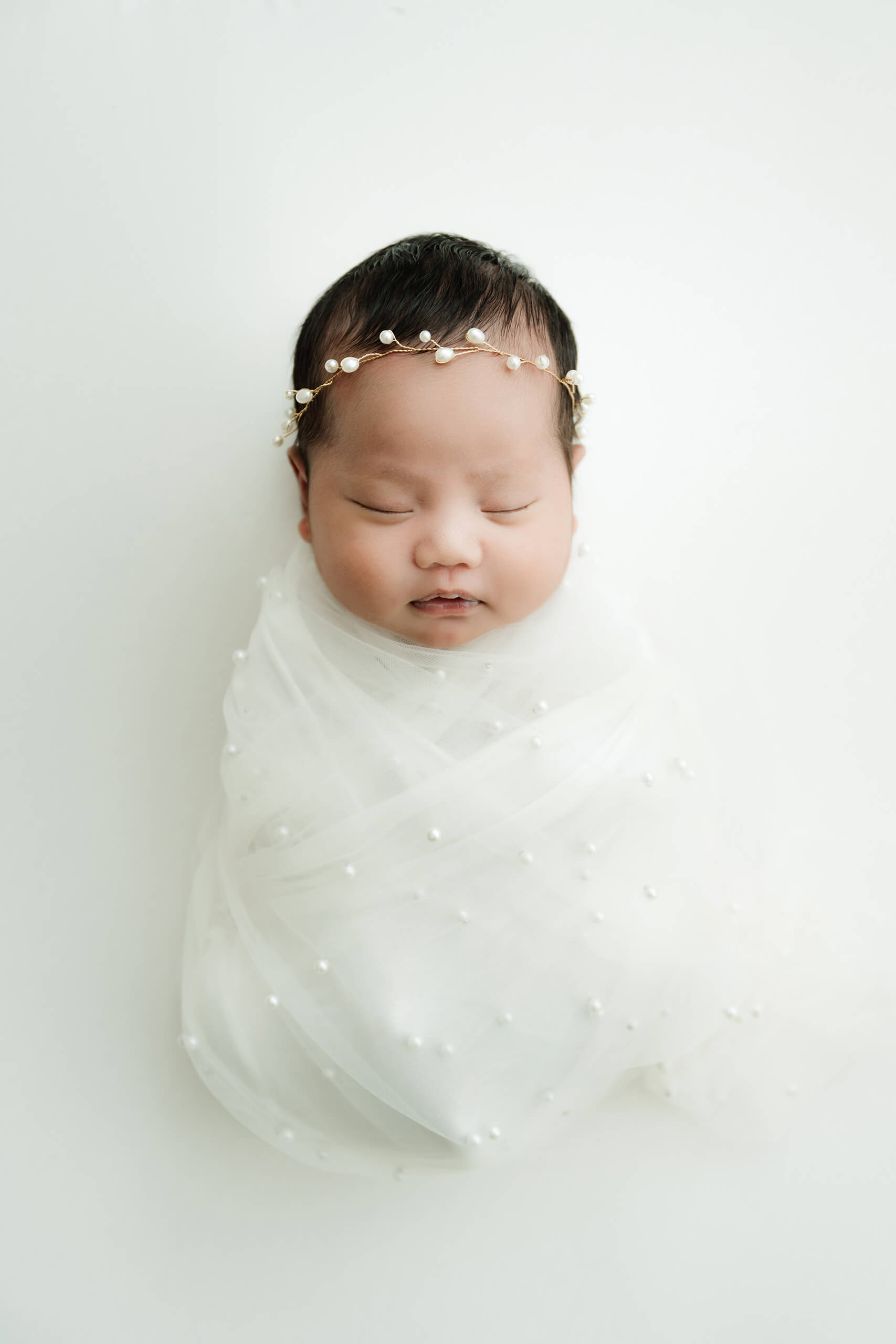 Sleeping newborn wrapped in sheer white fabric with pearl details and wearing a delicate pearl headband.