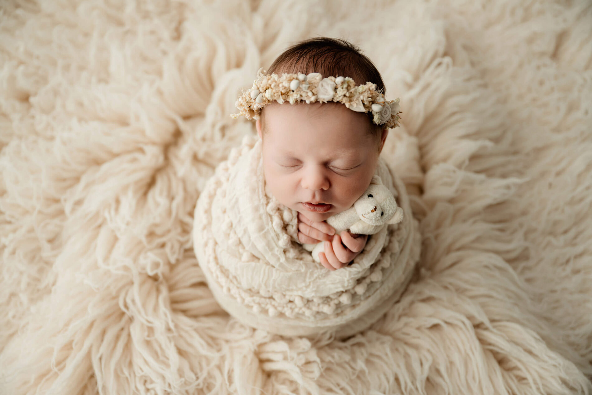 Sleeping newborn wrapped in cream textures, wearing a floral crown and holding a small white teddy bear.