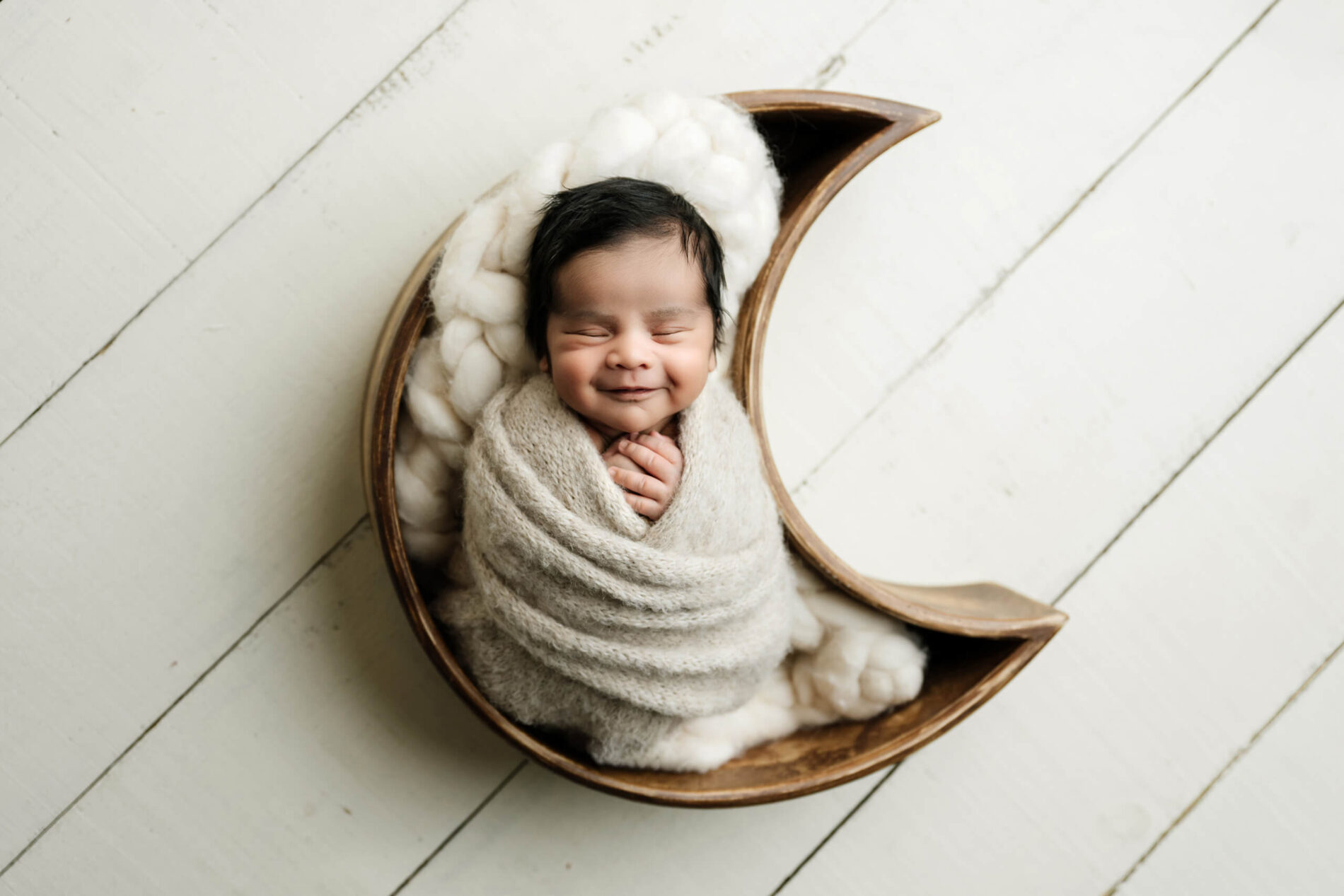 Smiling newborn wrapped in cream with hands beneath the chin, posed inside a wooden crescent moon prop.
