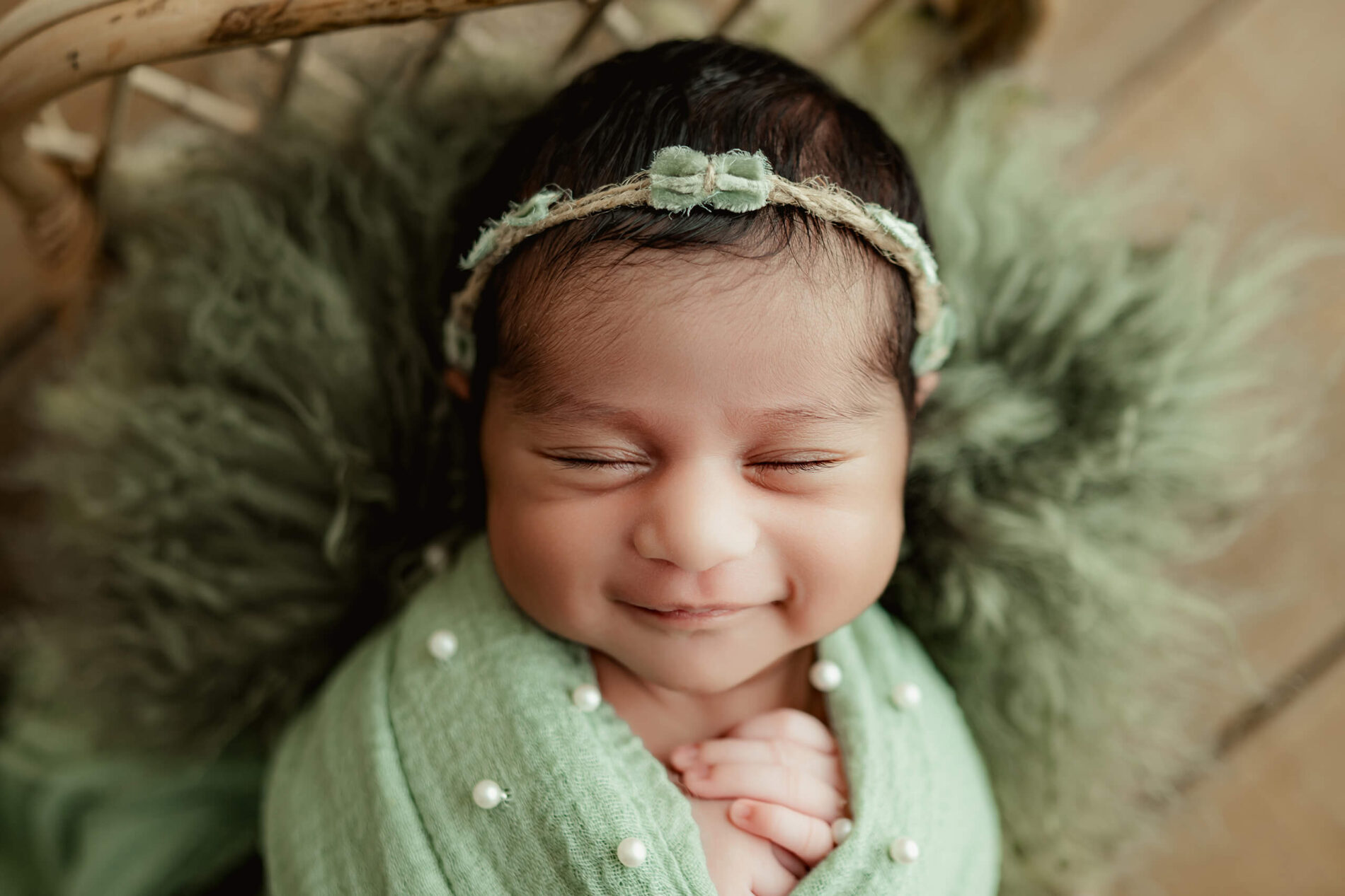 Smiling newborn wrapped in sage green with pearl details and wearing a small green bow headband.