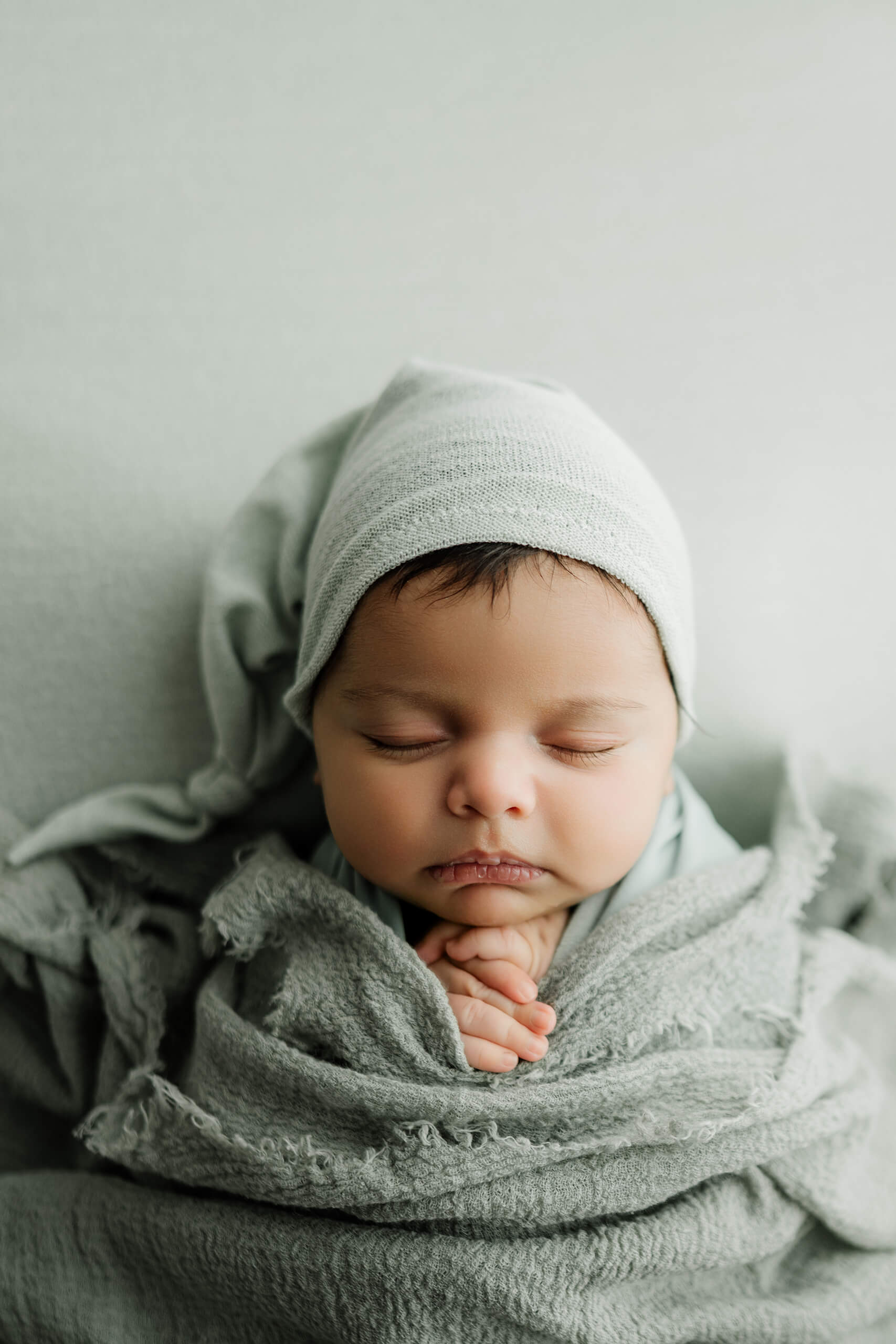 Sleeping newborn wrapped in soft sage-gray layers with a matching bonnet and hands tucked beneath the chin.