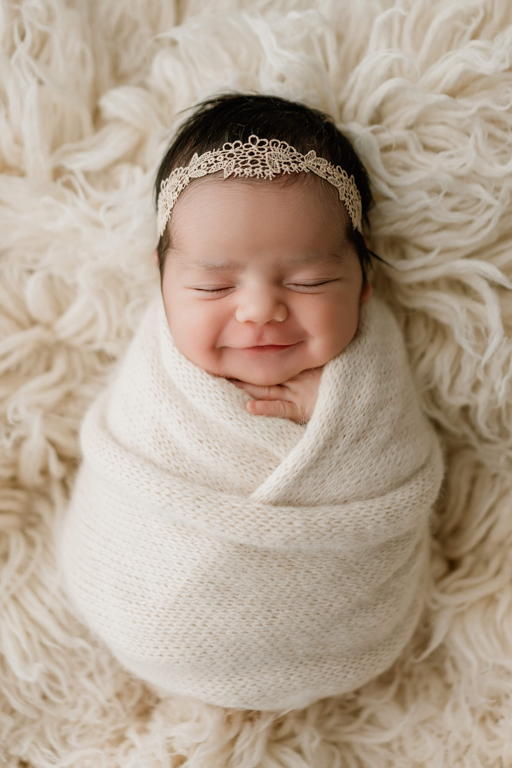 Smiling newborn wrapped in cream and wearing a delicate lace headband on a textured cream blanket.