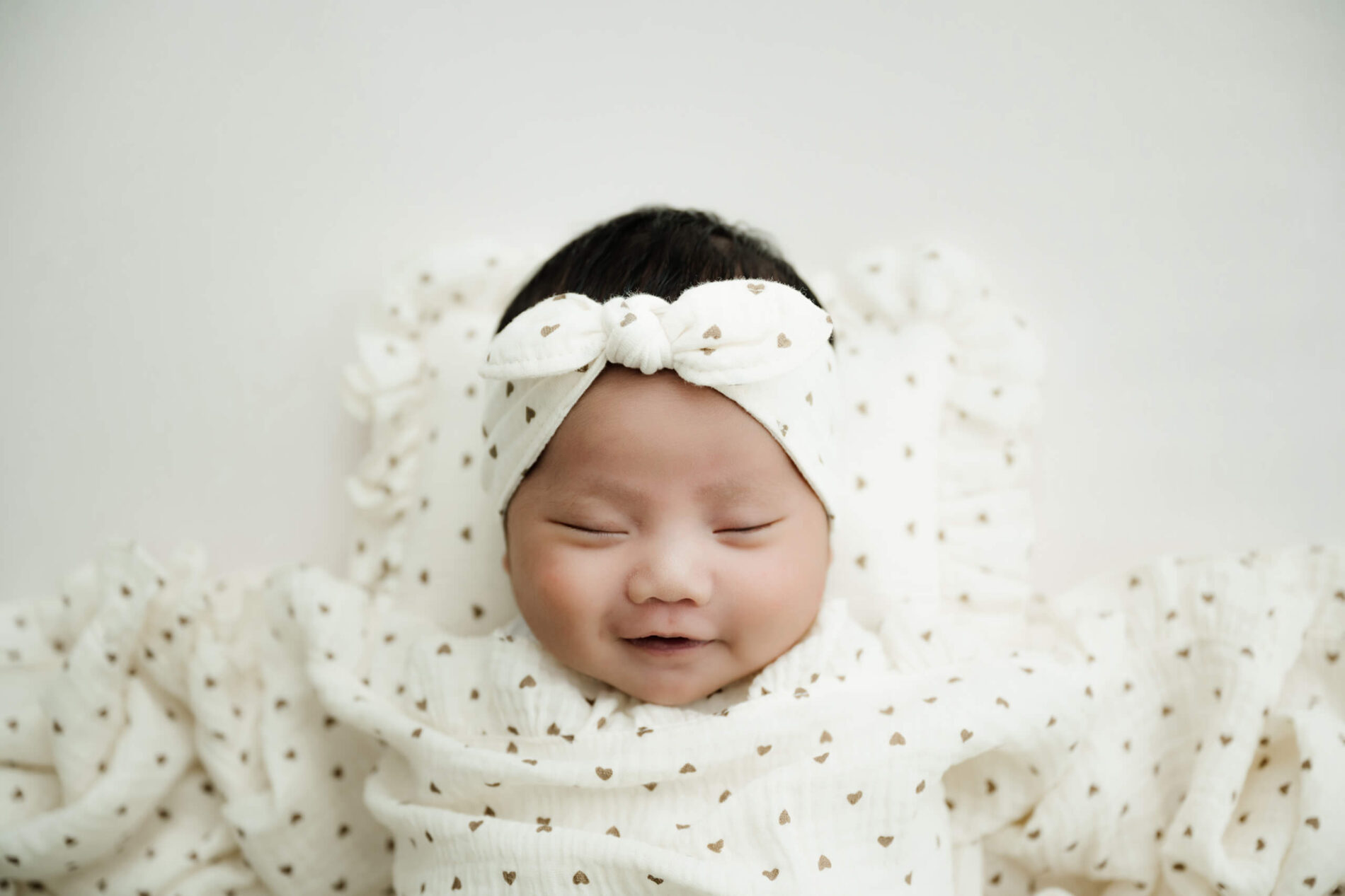 Smiling newborn wrapped in white fabric with tiny gold hearts and wearing a matching bow headband.