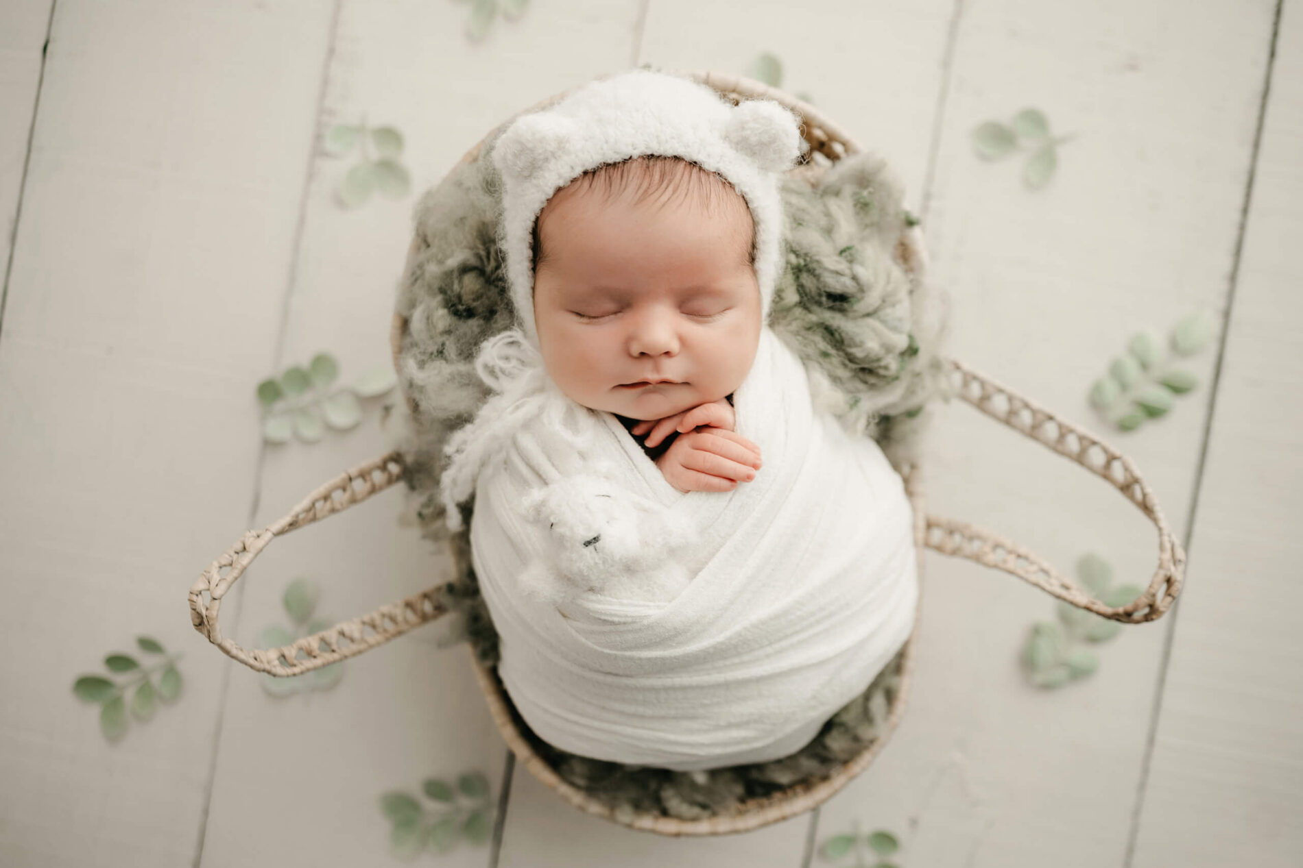 Sleeping newborn wrapped in white with a bear-ear bonnet, posed in a woven rabbit-shaped basket with eucalyptus leaves.