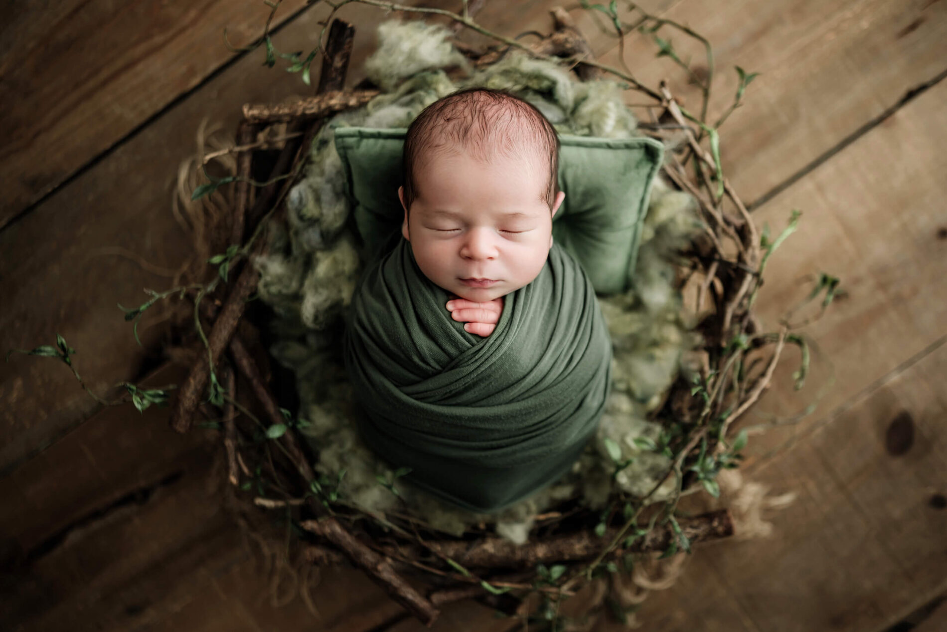 Sleeping newborn wrapped in deep green, posed with hands beneath the chin in a rustic twig nest with greenery.
