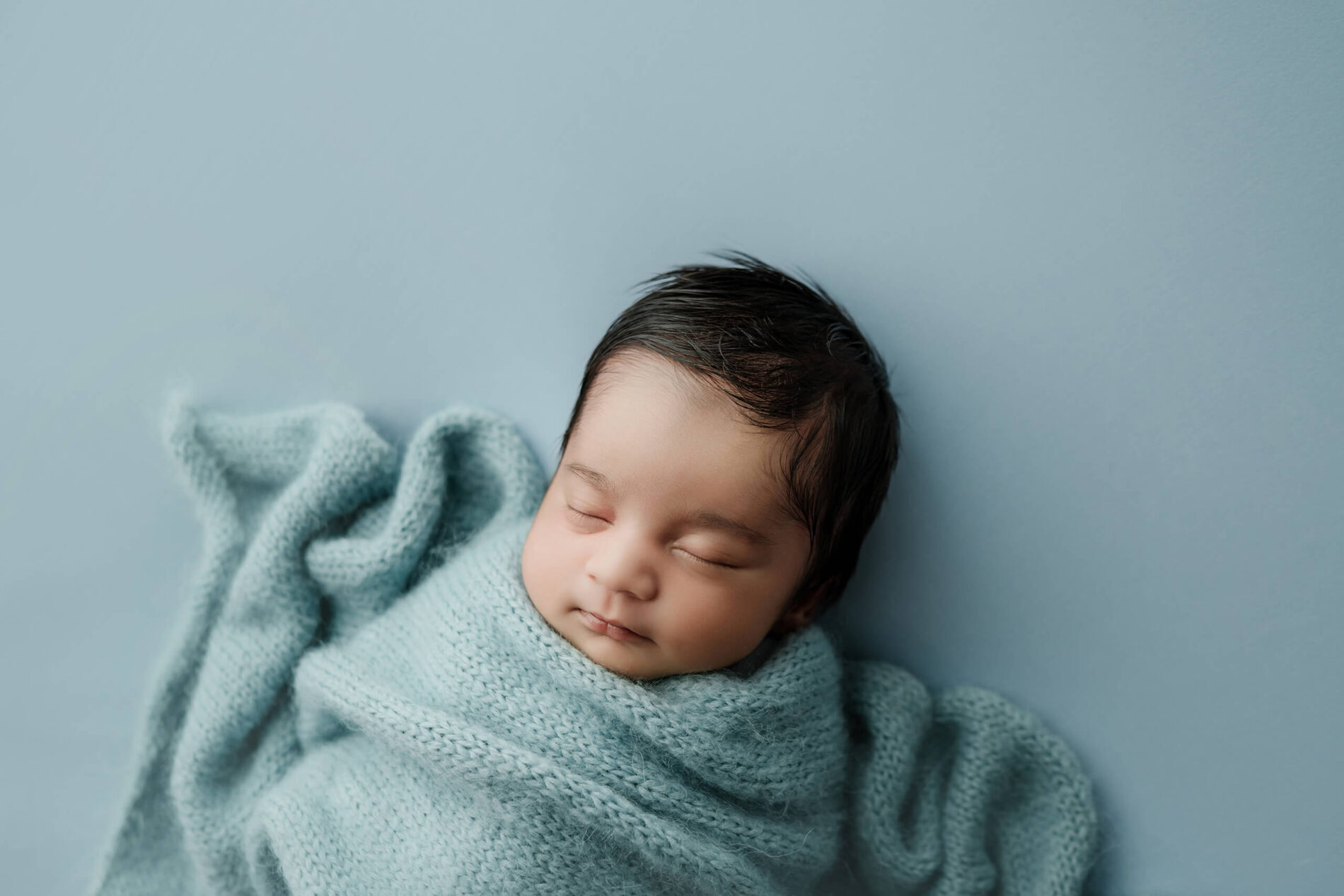 Sleeping newborn wrapped in a pale blue knit layer against a matching blue backdrop.