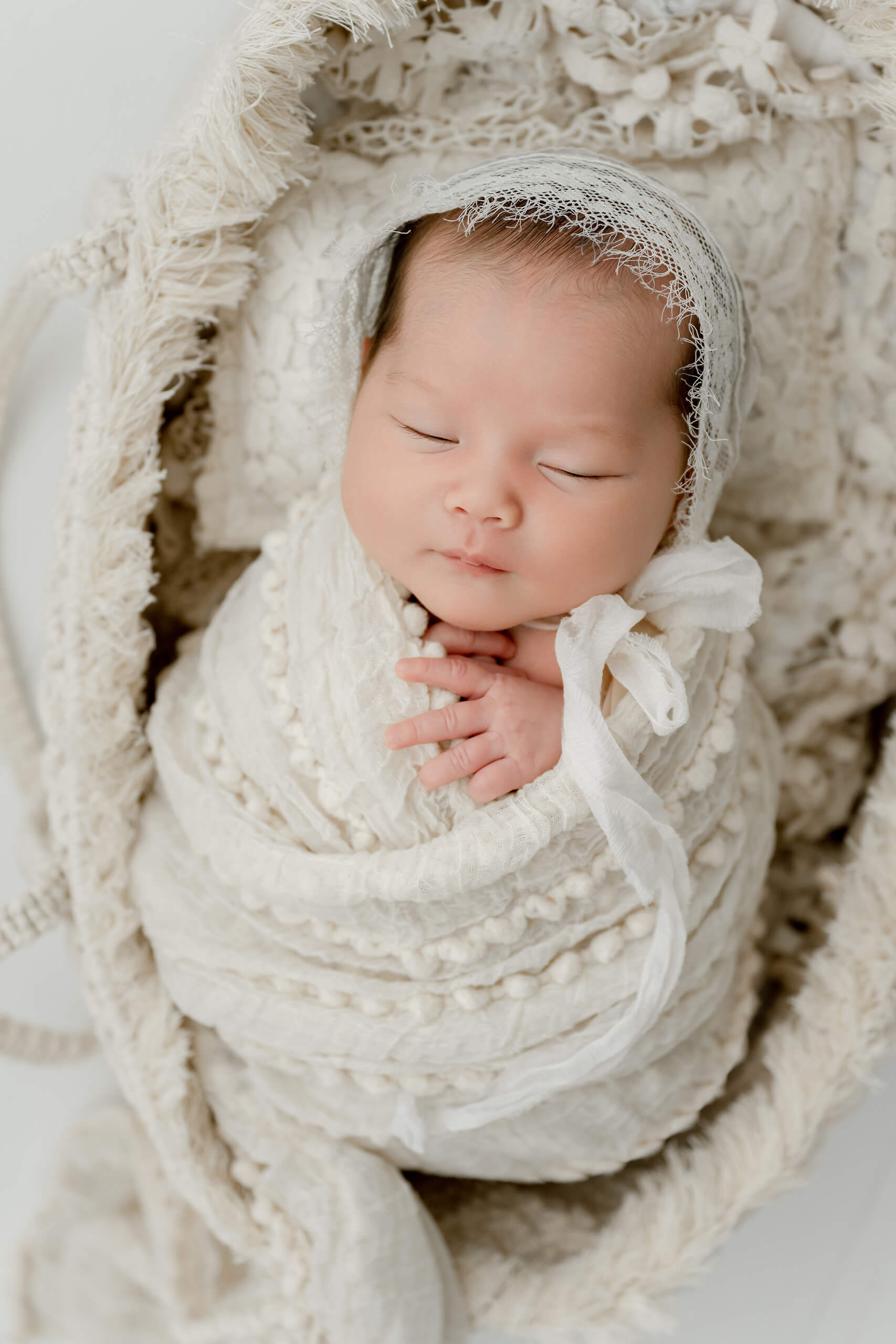 Sleeping newborn wrapped in white textured layers and wearing a delicate lace bonnet with a side bow.