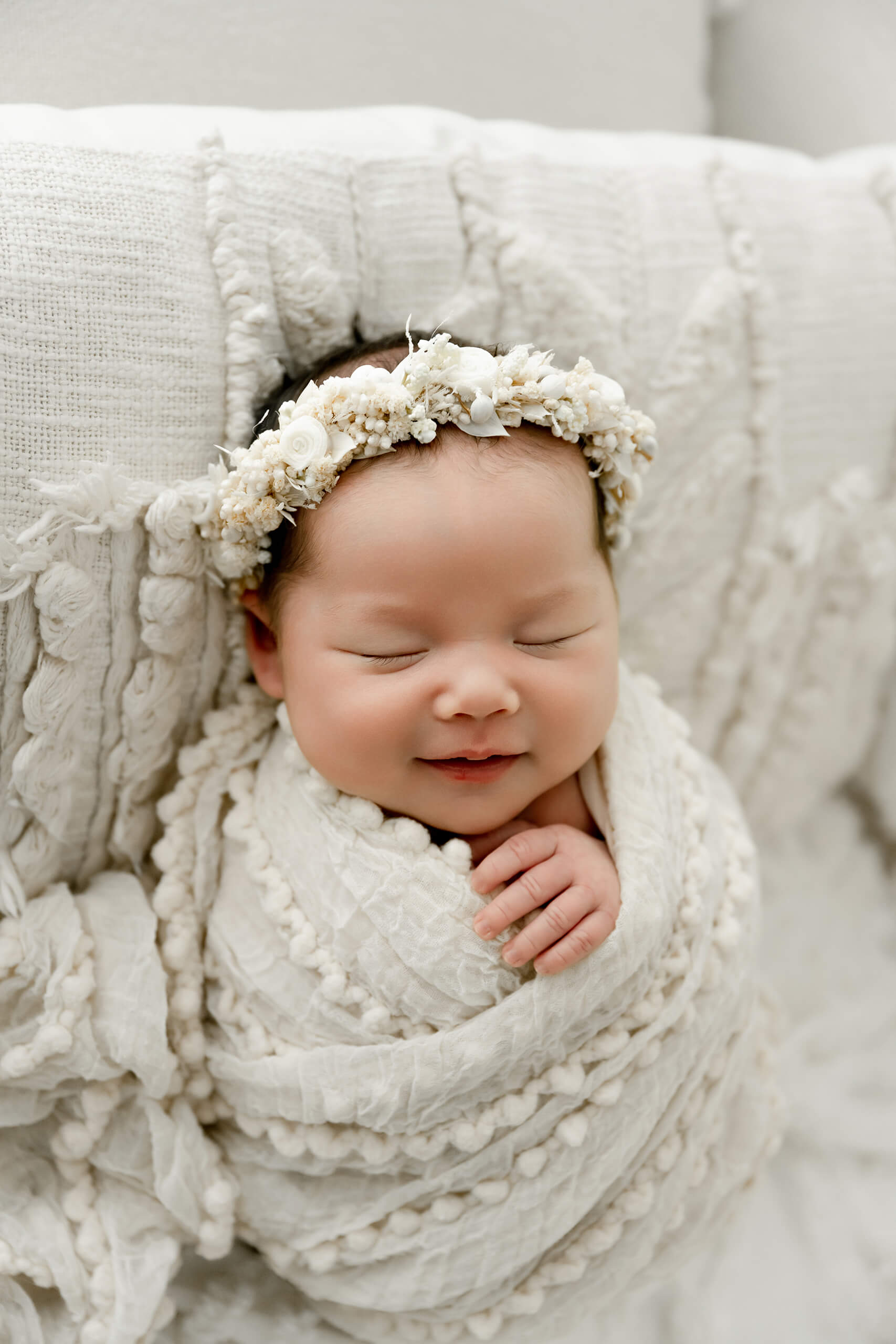 Smiling newborn wrapped in cream textured fabric and wearing a white floral crown.