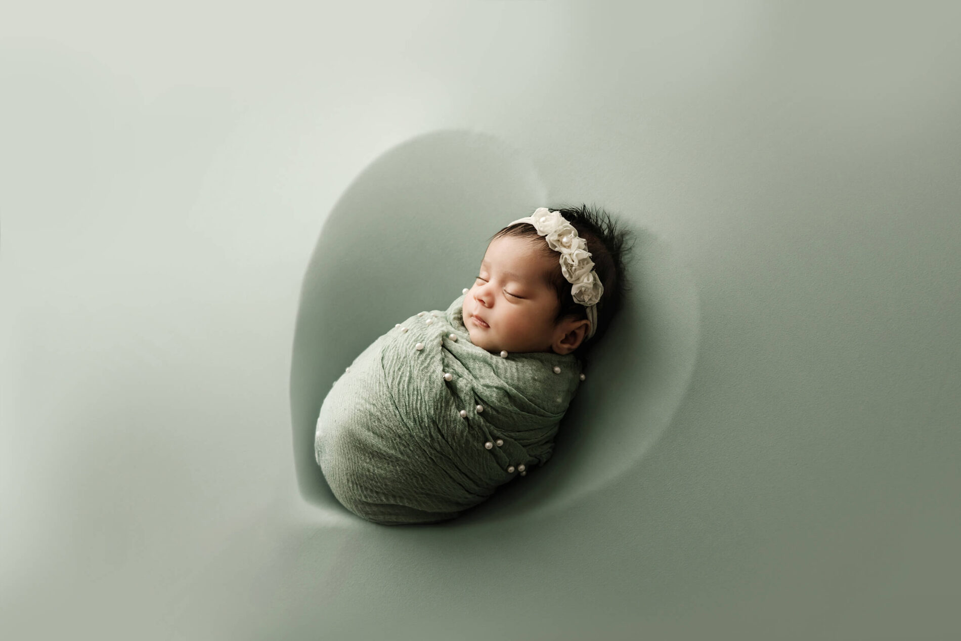 Sleeping newborn wrapped in sage green with pearl details and wearing a white floral headband on a matching backdrop.