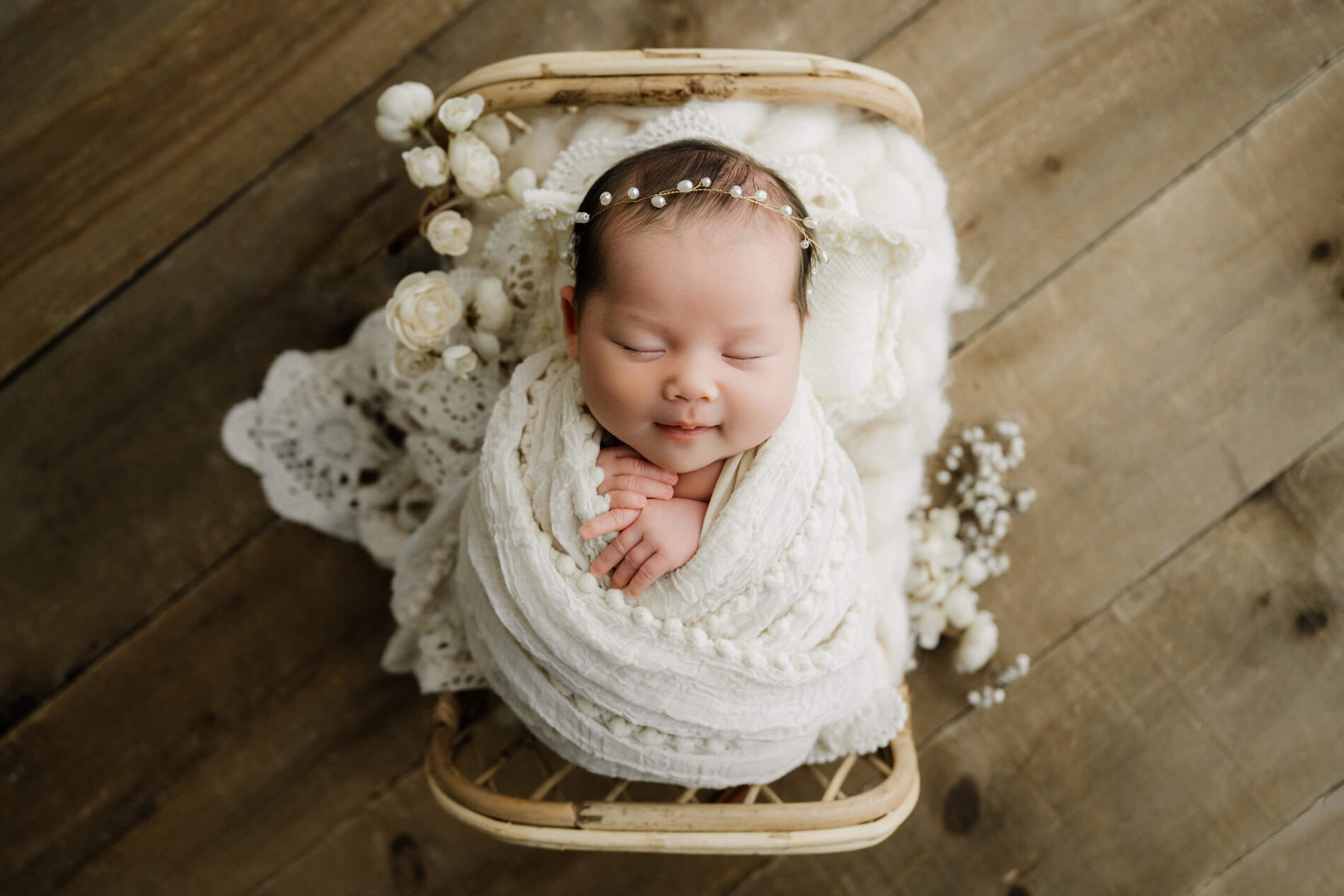 Sleeping newborn wrapped in white textured layers with a pearl headband, posed in a small rattan bed with cream flowers.
