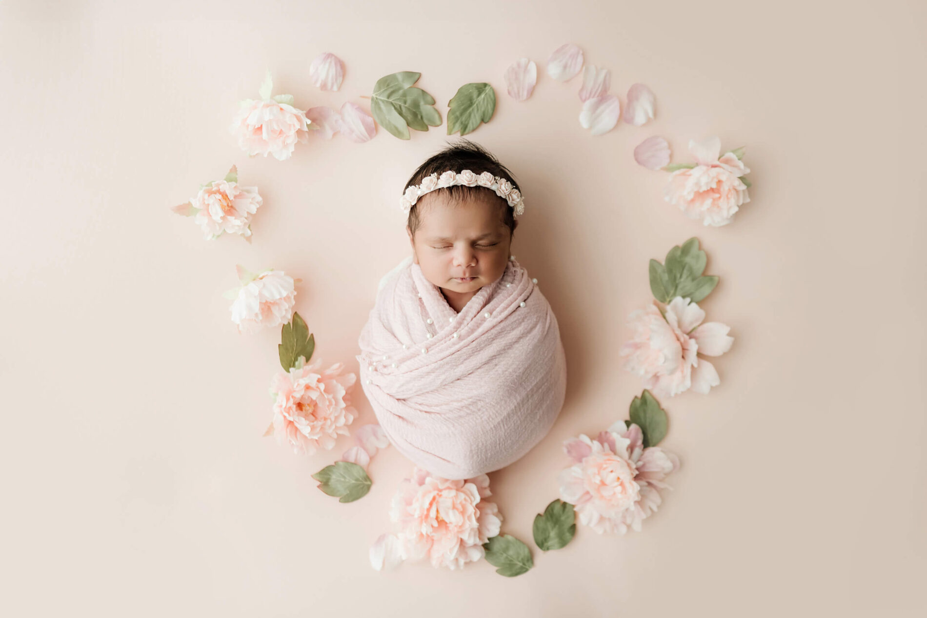 Sleeping newborn wrapped in blush pink with pearl details, surrounded by pink flowers and green leaves arranged in a heart.