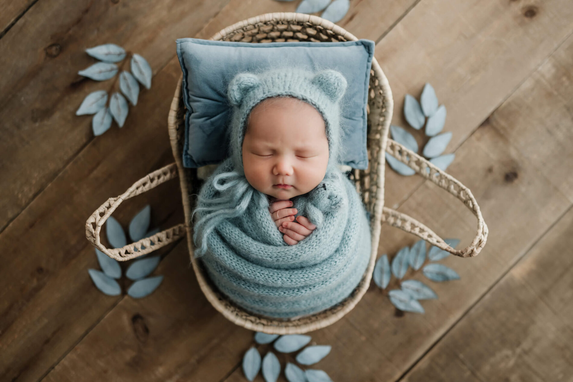 Sleeping newborn wrapped in dusty blue with a bear-ear bonnet and matching teddy bear, posed in a woven basket with blue leaves.
