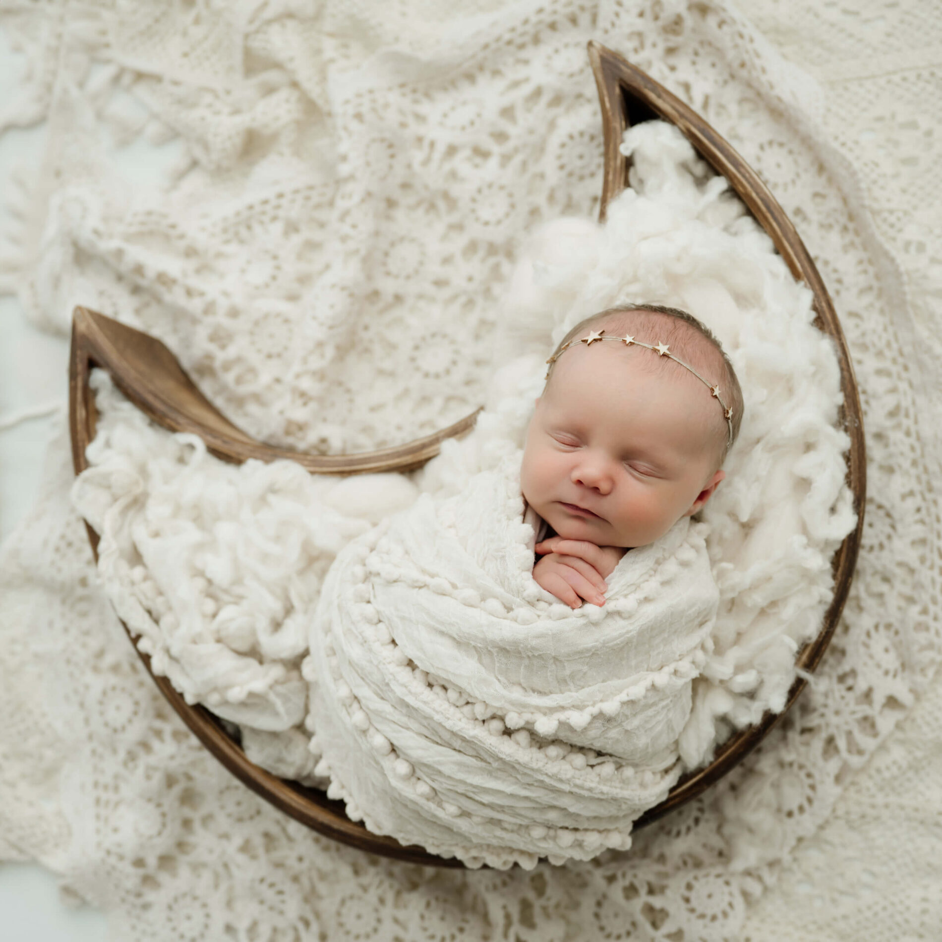 Sleeping newborn wrapped in white with a delicate star headband, posed in a wooden crescent moon prop on cream lace.