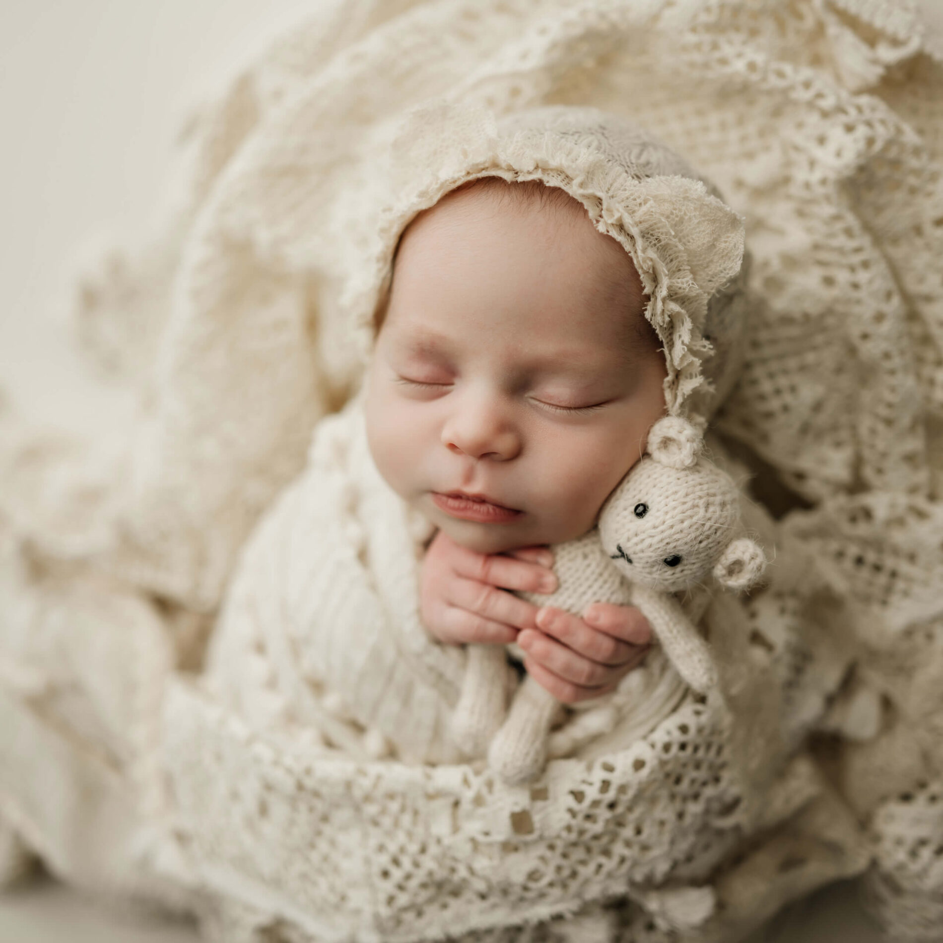 Sleeping newborn wearing a cream lace bonnet and cuddling a small knitted lamb among soft cream layers.