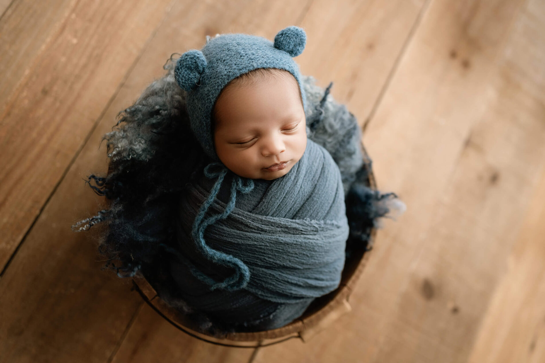 Sleeping newborn wrapped in blue-gray with a matching bear-ear bonnet, posed in a wooden bowl with dark textured wool.