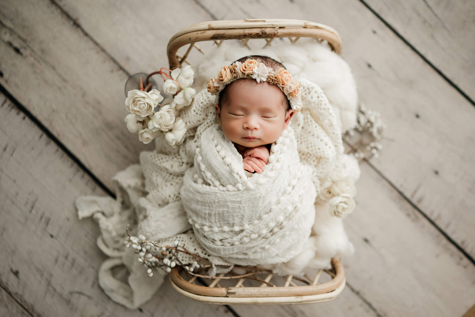 Sleeping newborn wrapped in white and wearing a peach floral crown, posed in a rattan bed with cream flowers.